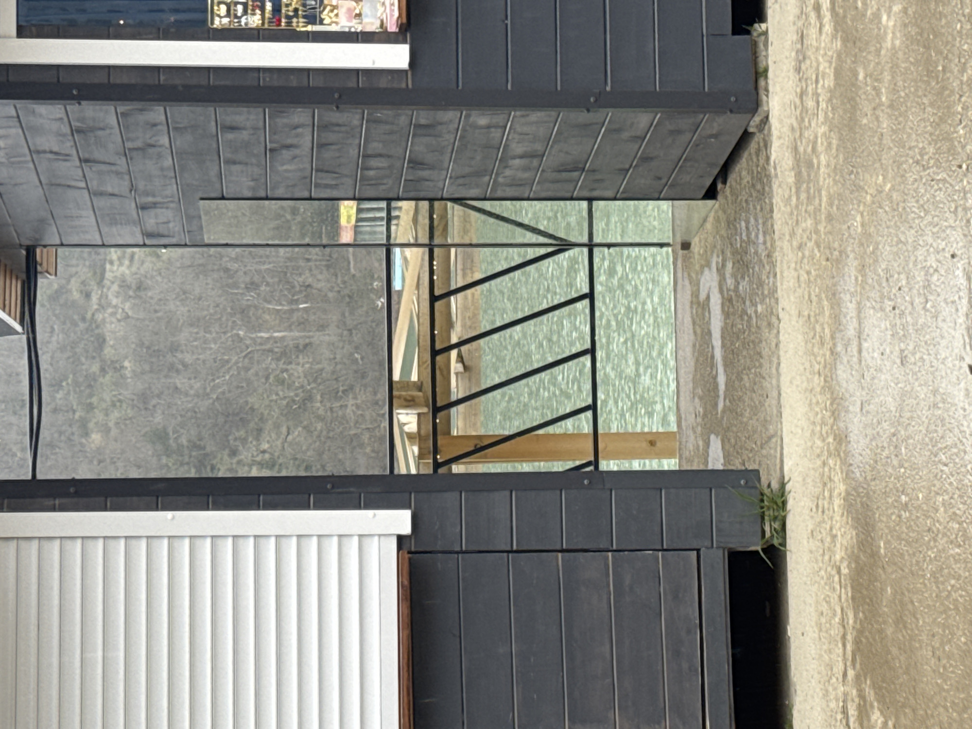 The image shows a partial view of two adjacent, dark-colored wooden structures, possibly sheds or small buildings, in Sukko, Russia. Between the structures, a glimpse of the outdoors is visible. There is a black metal railing and some wooden supports, suggesting a deck or similar outdoor space. Reflected in the glass in the wooden structure on the left is a forested area and light-colored building across the water. To the right of the dark structure is a light colored wall. Some vegetation is visible along the bottom right of the image.