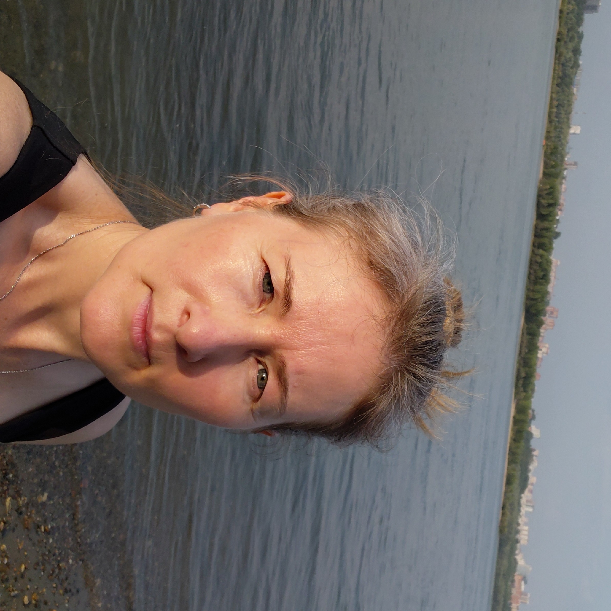 A woman with blonde hair pulled back in a bun is taking a selfie outdoors. She is wearing a black top and a silver necklace. The background shows a body of water with a city skyline and trees on the opposite shore. This appears to be the Ob River in Novosibirsk, Russia, possibly near the embankment or the Zoo Bridge.