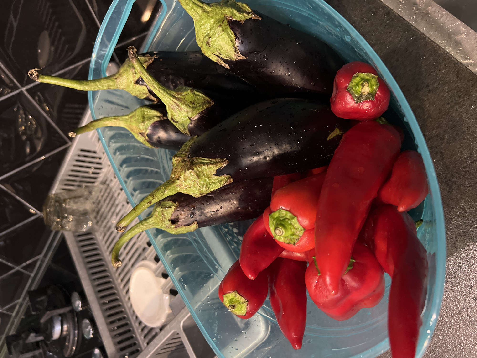 A close-up, overhead shot shows a blue plastic basket filled with fresh produce, including several dark purple eggplants and a variety of red peppers. The eggplants are plump with green stems, while the peppers range from long and slender to small and round. The vegetables appear to have been recently washed, with visible water droplets on their surfaces. The basket is placed on a dark, speckled countertop, and in the background, a stainless steel dish rack with a glass and a white lid can be seen next to a stovetop. The scene is indoors, likely a kitchen, given the presence of kitchen appliances and a dish rack. The image does not contain any information related to the city of Tsodoreti, Georgia.