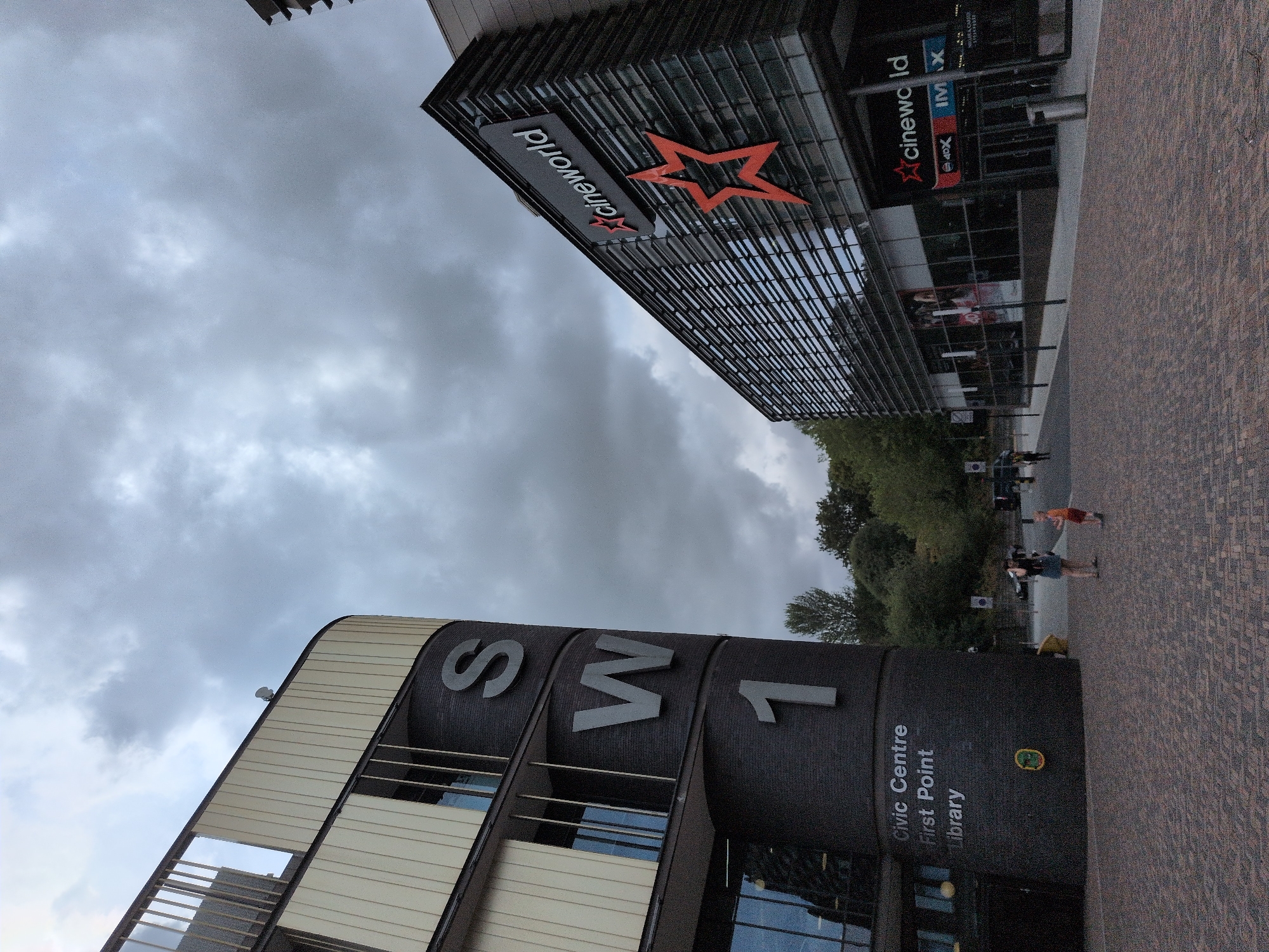 This image depicts the exterior of the civic centre and a Cineworld cinema, located in Telford, United Kingdom. The civic centre, on the left, is a multi-story building with a curved facade. Large grey letters spelling "SW 1" are prominently displayed on the dark brickwork of the building's cylindrical section. The text "Civic Centre First Point Library" is also visible on the lower portion of this building.
To the right, the Cineworld cinema is a modern building with a glass facade featuring horizontal louvers. A prominent red star logo and the "cineworld" text are visible on its exterior, along with smaller signs indicating "4DX" and "IMAX". The entrance to the cinema is visible, with people in the foreground walking across a brick-paved plaza.
The sky is overcast with heavy grey clouds. Trees and greenery are visible in the background, suggesting an urban setting with some natural elements. The overall impression is of a public gathering space with entertainment and civic facilities.