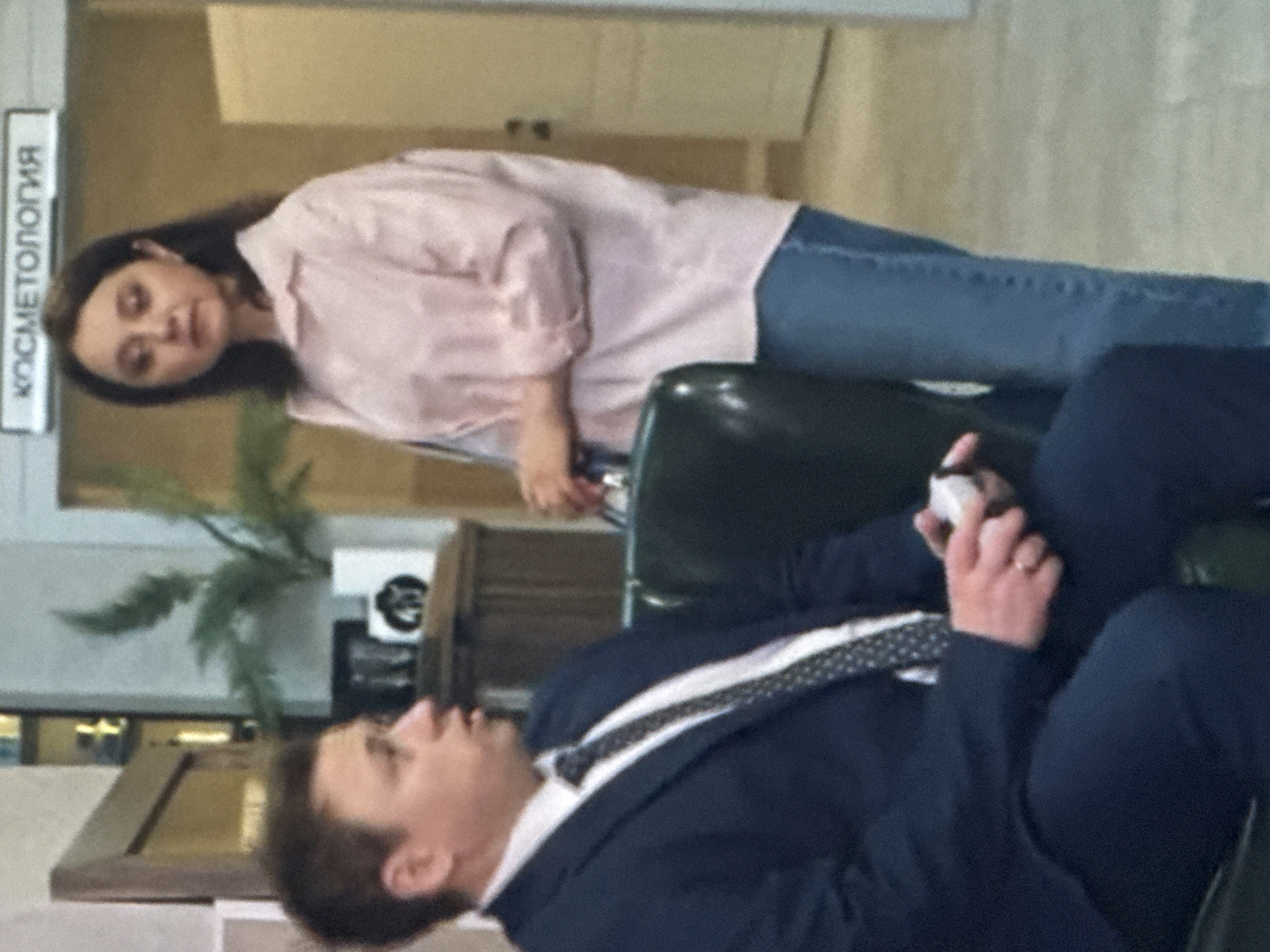 The image shows two people in an indoor setting, possibly an office or waiting area. The person in the foreground is wearing a dark blue suit and white shirt with a patterned tie. They are seated and looking upwards. Their right hand is raised, holding a small white object, and a ring is visible on their left ring finger.
In the background, another person is visible. They are wearing a light pink collared shirt and blue jeans. Their hair is dark brown. They are also seated and looking towards the viewer. Next to them, on the wall, is a white sign with black text. The text on the sign reads "КОСМЕТОЛОГИЯ", which is Russian for "Cosmetology".
The scene appears to be indoors with neutral-colored walls. A plant is visible to the left of the person in the background. The lighting suggests it is daytime. The overall mood is somewhat casual and unposed. There are no clear indications of the time of day or weather conditions outside, but the indoor setting is well-lit. The presence of the "Cosmetology" sign suggests the location might be a salon or a medical facility specializing in cosmetic treatments.