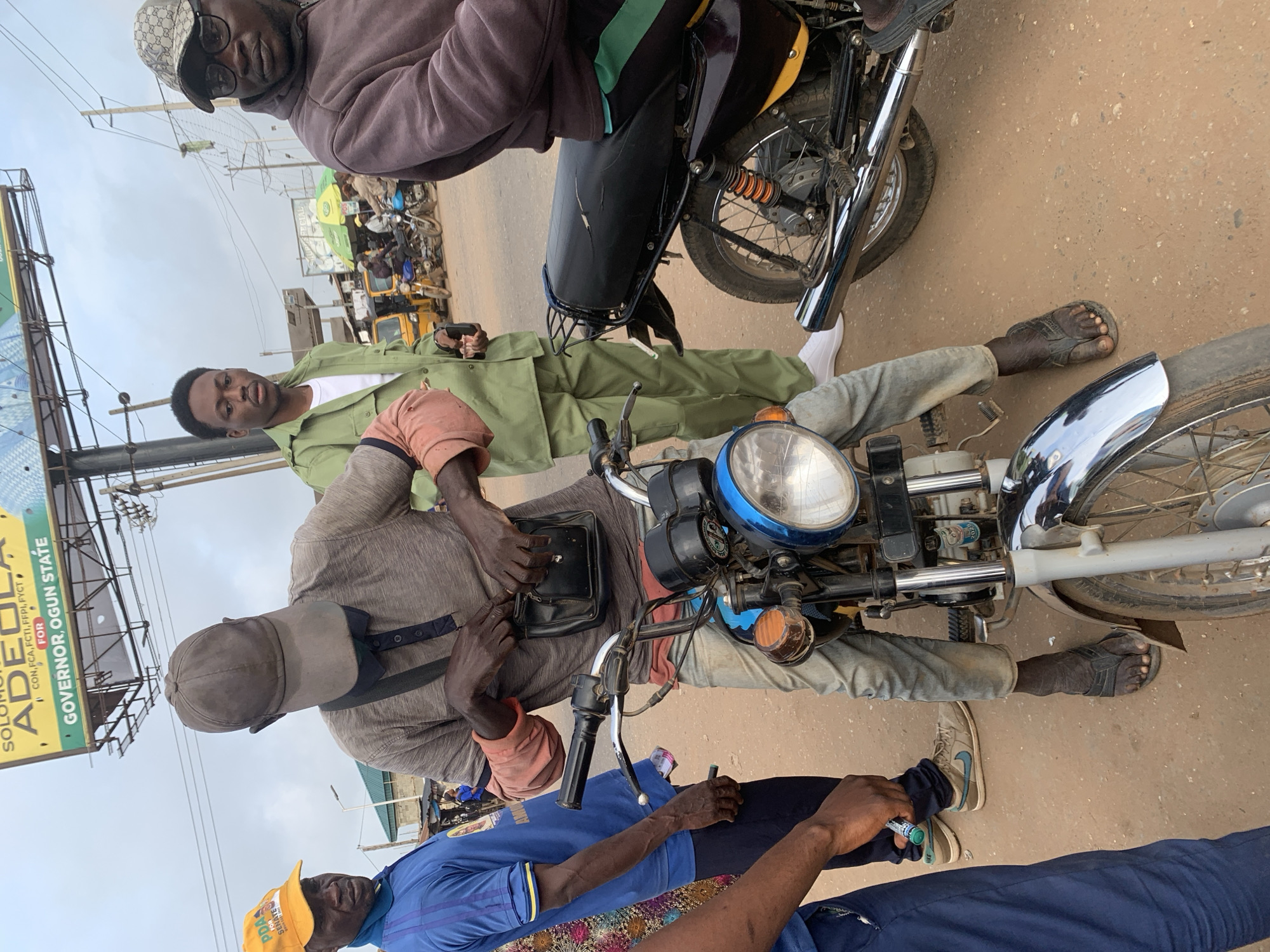 In Shagamu, Nigeria, a group of men are gathered on a dirt road. A man wearing a grey t-shirt, light blue jeans, and black sandals sits on a motorcycle. He is reaching into a black pouch hanging from his shoulder. Another man, to the left, wears a blue t-shirt and a yellow cap with "PDP FOR SENATE" printed on it. A third man, in a green uniform and white t-shirt, stands to the right of the motorcycle rider, holding a small object in his hand. A fourth man, wearing a grey hooded sweatshirt and a cap with a pattern, is seated on another motorcycle in the background. The environment appears to be a busy street with other motorcycles and tricycles visible. A large billboard with "SOLOMON ADEOLA FOR GOVERNOR, OGUN STATE" is prominently displayed in the background.