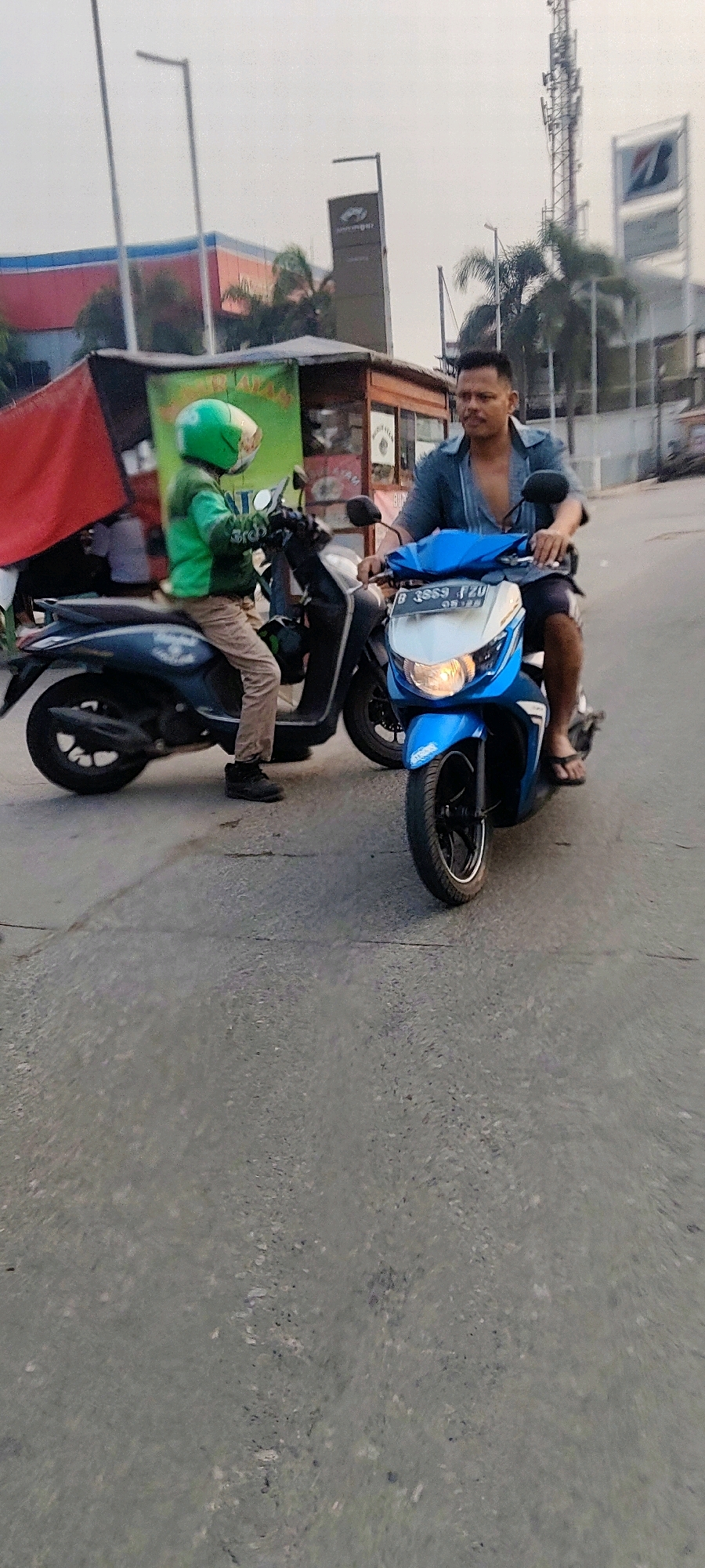 Two men are on motorcycles on a paved road. The man on the left is wearing a green helmet and jacket, brown pants, and black shoes, and is on a dark blue scooter. The man on the right is wearing a blue patterned shirt and dark shorts with flip-flops, and is on a blue scooter. Both men appear to be looking forward, and the scooters are stopped side-by-side on the road. In the background, there is a building with a red and blue facade, a cell tower, and palm trees. There are also some food stalls visible. The sky is overcast. The scene is likely in the city of Mekarmukti, Indonesia, given the presence of the food stalls and the style of the buildings.