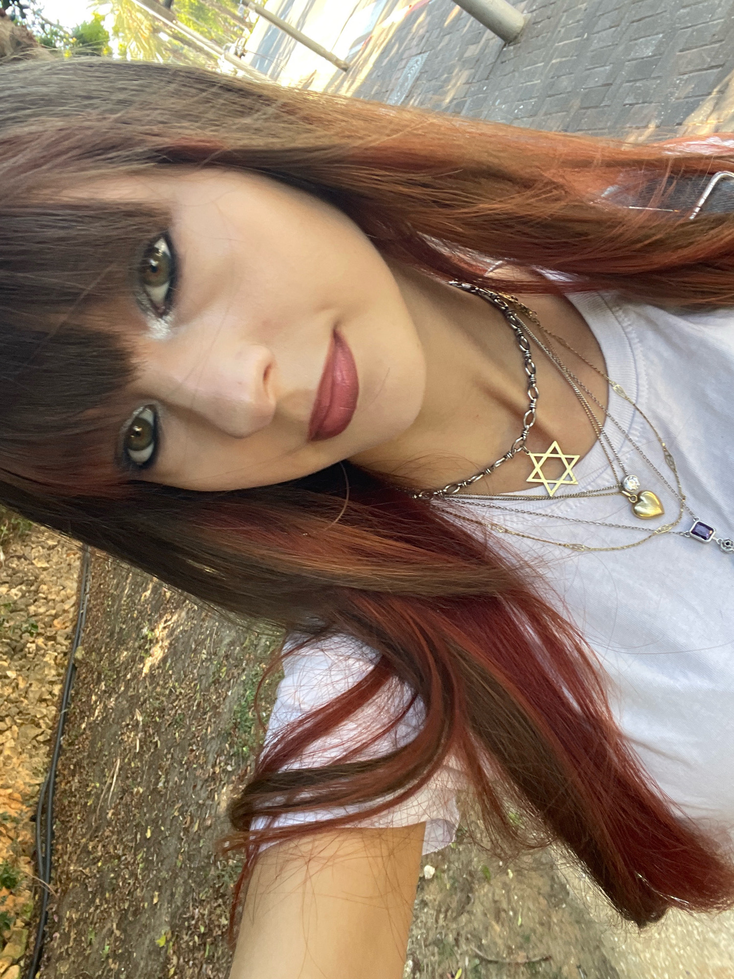 A young woman with brown hair streaked with red is taking a selfie outdoors in Raanana, Israel. She is wearing a white t-shirt and multiple necklaces, one of which features a Star of David pendant. The background shows a paved walkway and some greenery, consistent with a park or street environment in Raanana. The image is shot from a low angle, with the woman's face tilted towards the camera.