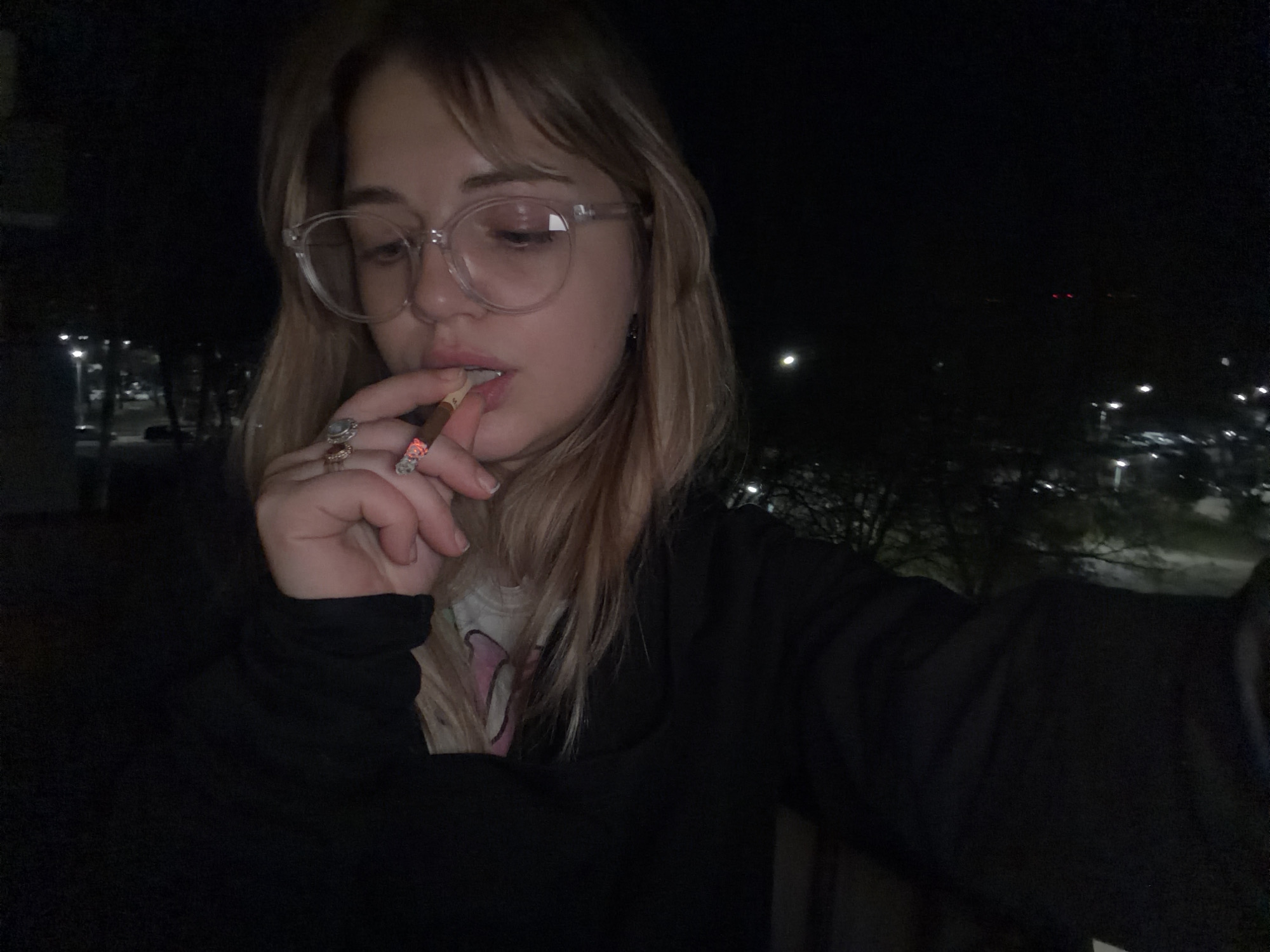 A person with blond hair and wearing clear-framed glasses holds a lit cigarette to their lips. They are wearing a dark-colored sweater over a t-shirt. The location appears to be outdoors at night, with lights visible in the background. There is no definitive information in the image to confirm the location is Naberezhnye Chelny, Russia.