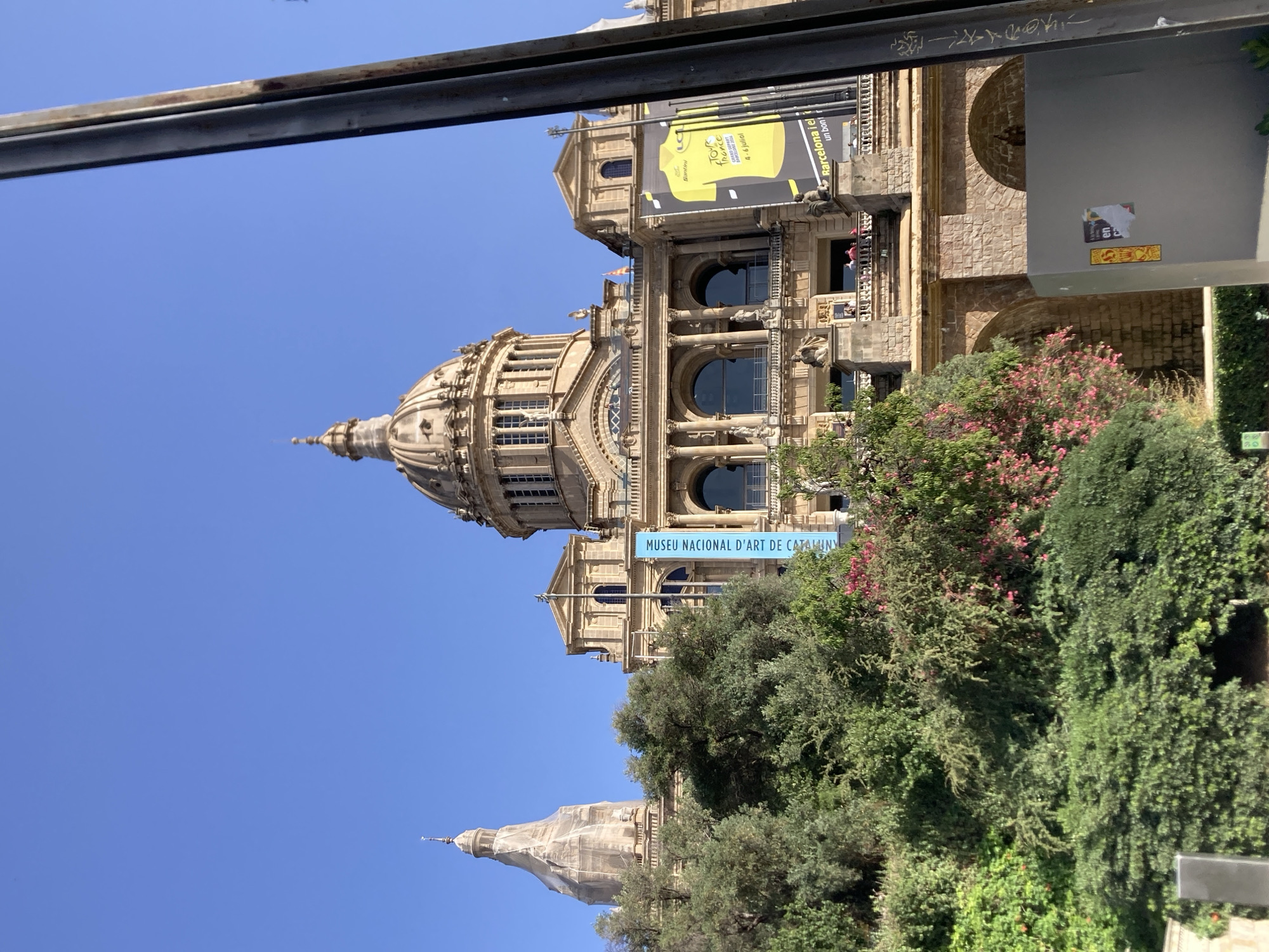 The image captures a sunny day in Barcelona, Spain, with the prominent facade of the Museu Nacional d'Art de Catalunya (MNAC) as the central subject. The MNAC is a grand building with multiple domes and classical architectural elements, including columns and arched windows. A large banner advertising the "Tour de France" event, with a yellow cycling jersey graphic and Spanish text, hangs from the building. Another blue banner with text in Catalan, "MUSEU NACIONAL D'ART DE CATALUNYA," is also visible. Lush green trees and some flowering bushes are in the foreground, partially obscuring the lower levels of the building. The sky is a clear, bright blue. A tall, dark vertical pole, possibly part of a structure or scaffolding, partially obscures the right side of the image. There are a few people visible on a balcony on the MNAC's second level.