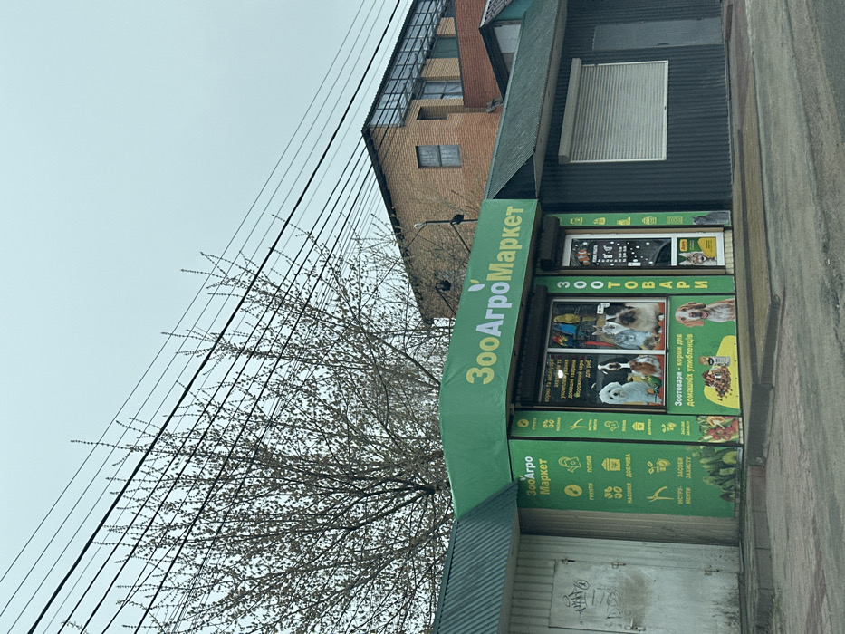 The image shows a pet supply store called "ZooAgroMarket" located in Tarasivka, Ukraine, at coordinates 50.343, 30.304. The store has a bright green facade with a large sign featuring the store name in bold, white lettering. The storefront windows display various pet products and supplies, including pet food, toys, and accessories. The windows also feature images of several dogs, cats, and birds. There are several power lines and a large tree in the foreground, and a brick building is visible behind the store. The pavement in front of the store appears to be made of asphalt.