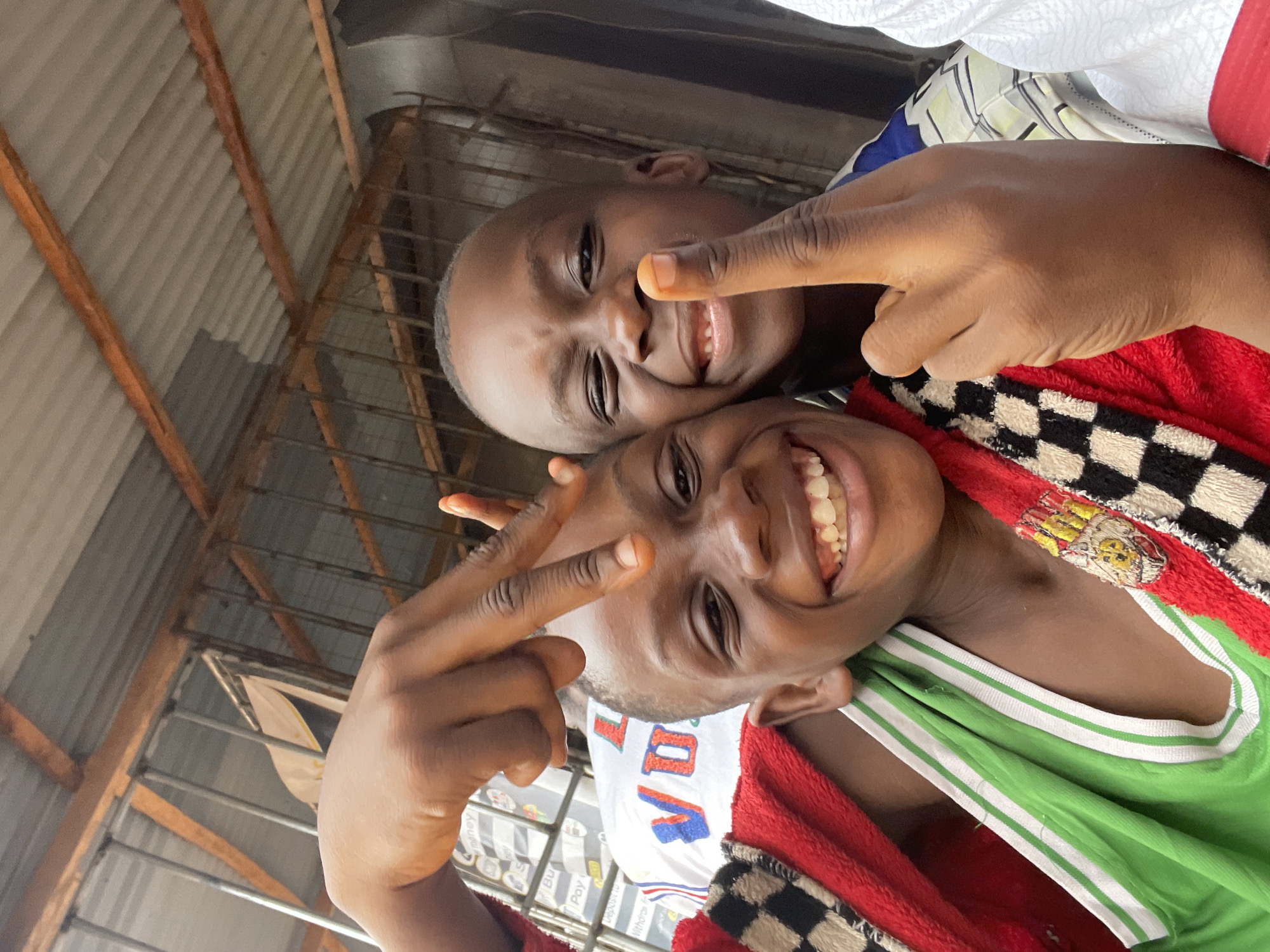Two smiling boys are photographed closely in what appears to be Onitsha, Nigeria. One boy is making a peace sign, while the other is pointing towards the camera. The environment includes corrugated metal roofing and metal framework suggesting an open-air market stall or similar commercial structure common in Onitsha. The boy at the bottom is wearing a green shirt with white stripes and is covered with a red blanket, and the boy above is wearing a blue and white checkered top.