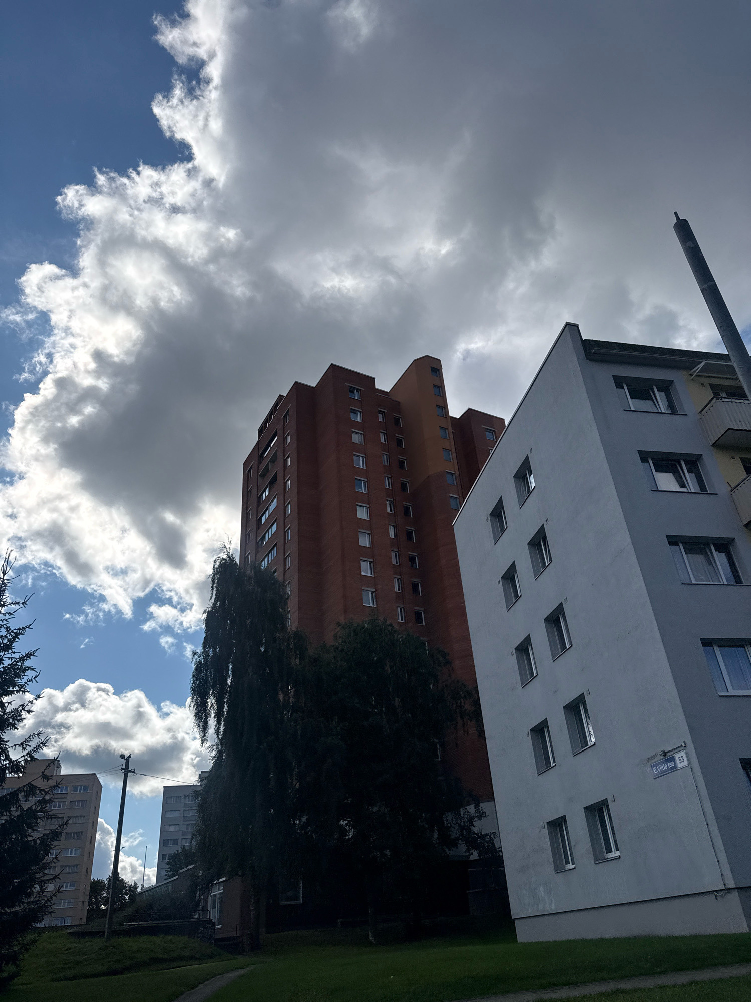 The image captures a street view in Tallinn, Estonia, likely from the Lasnamäe district, known for its Soviet-era apartment blocks. In the foreground, a light blue, angular apartment building is prominently featured, its facade punctuated by numerous windows. To its left, a taller, reddish-brown brick building, characteristic of the area's architecture, rises high into the sky. The sky is a dramatic mix of blue patches and heavy, grey clouds, suggesting an overcast or transitional weather condition. A tall, dark tree with drooping branches partially obscures the view between the two buildings. In the background, other apartment buildings and a utility pole can be seen. A street sign on the light blue building reads "E. Vilde tee 53," indicating the specific location. The ground is covered with green grass, with a paved path visible. The image is taken from a low angle, emphasizing the height of the buildings and the expanse of the sky. No people are visible in the frame.