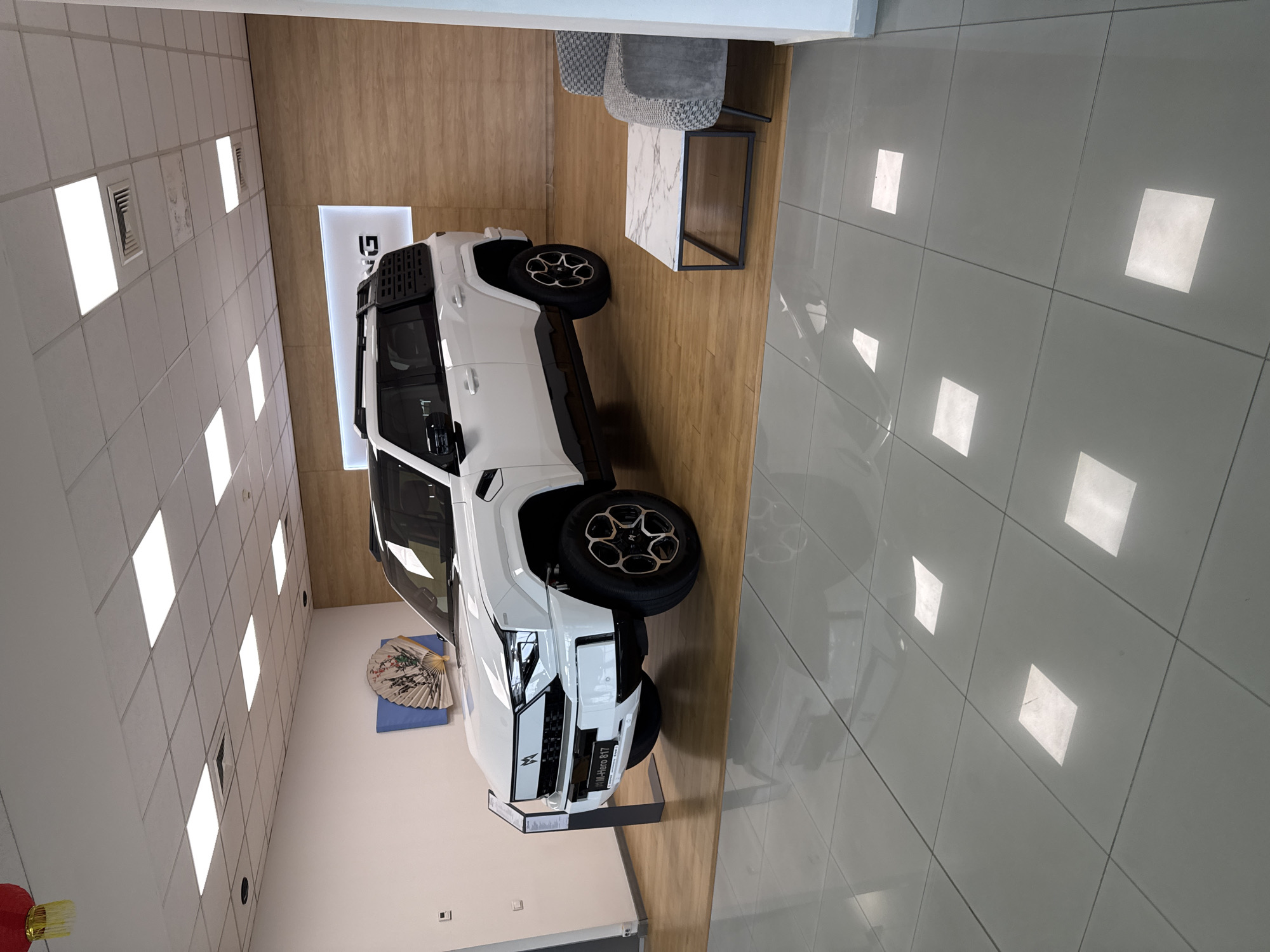 The image shows the interior of what appears to be a car dealership or showroom. A white vehicle, identified by the license plate as "WM-Hero 817," is prominently displayed. Other notable items include a table with chairs, a decorative fan on the wall, and a backdrop featuring graphics. Based on the license plate's likely origin, the location is suspected to be in Sofia, Bulgaria. The environment is well-lit with reflective tile flooring. There are no people visible.