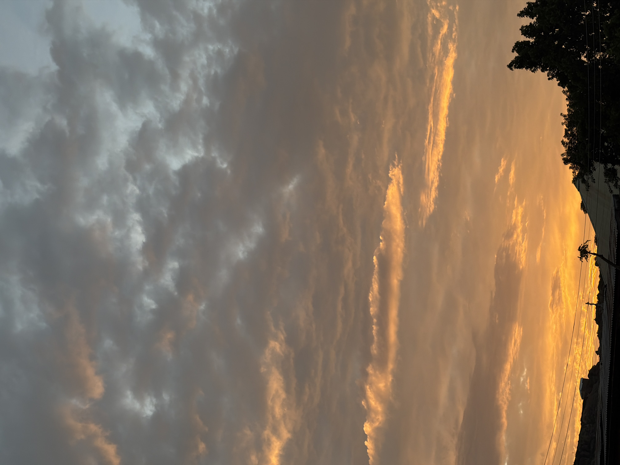 The image captures a dramatic sunset over Jos, Nigeria. The sky is a canvas of contrasting colors, with the left side dominated by dark, turbulent clouds that appear heavy and foreboding. As the eye moves to the right, the clouds transition into wispy streaks illuminated by the setting sun, casting a warm, golden glow. These illuminated clouds create a striking visual contrast with the darker ones, suggesting a dynamic and evolving weather pattern.
The horizon is silhouetted by dark shapes, hinting at the landscape below. On the far right, the dense foliage of a tree, likely native to the region, stands out against the vibrant sky. Beneath the tree, the edge of a building or structure is visible, along with what appear to be utility lines, suggesting an urban or semi-urban setting within Jos.
The time of day is clearly sunset, indicated by the intense golden and orange hues in the sky. The weather appears to be a mix of cloud cover, with some areas clear enough to allow the sunlight to break through and illuminate the clouds dramatically. The overall mood evoked by the image is one of natural beauty and perhaps a sense of transition or change, with the interplay of light and shadow creating a powerful visual statement. No text is visible in the image.