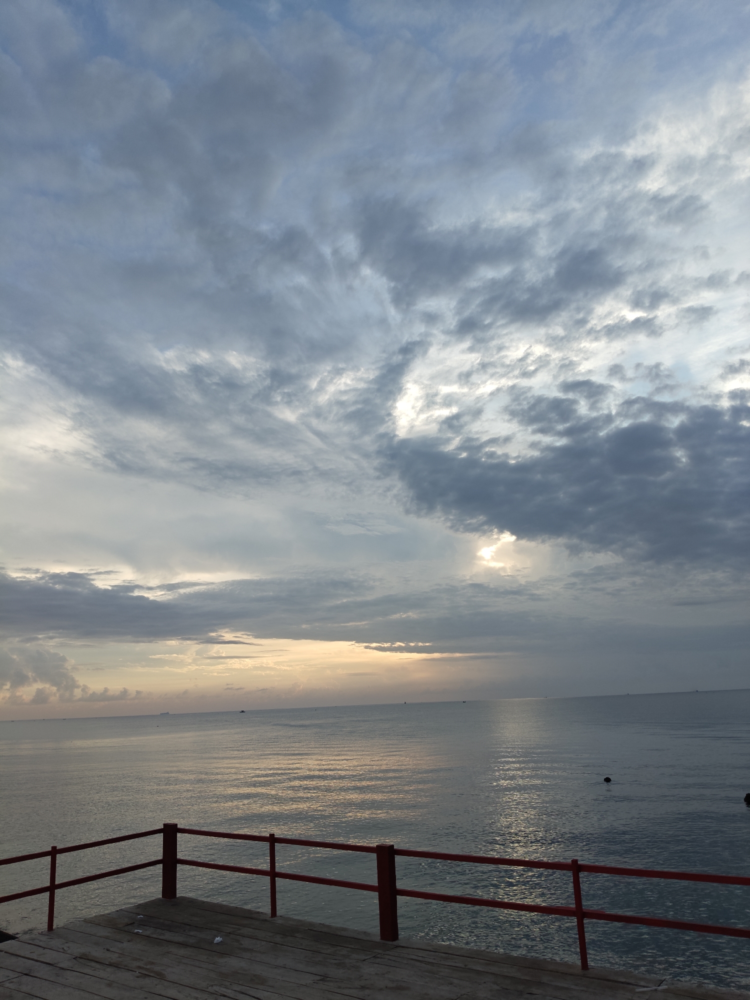 The image depicts a calm seascape with a wooden pier in the foreground, featuring red railings. The sky is filled with scattered clouds, and the sun is partially visible, casting a warm glow on the water. The ocean is serene, with gentle ripples reflecting the light. The overall atmosphere is tranquil, suggesting a peaceful morning or evening setting. The location is identified as Lhokseumawe, Indonesia.