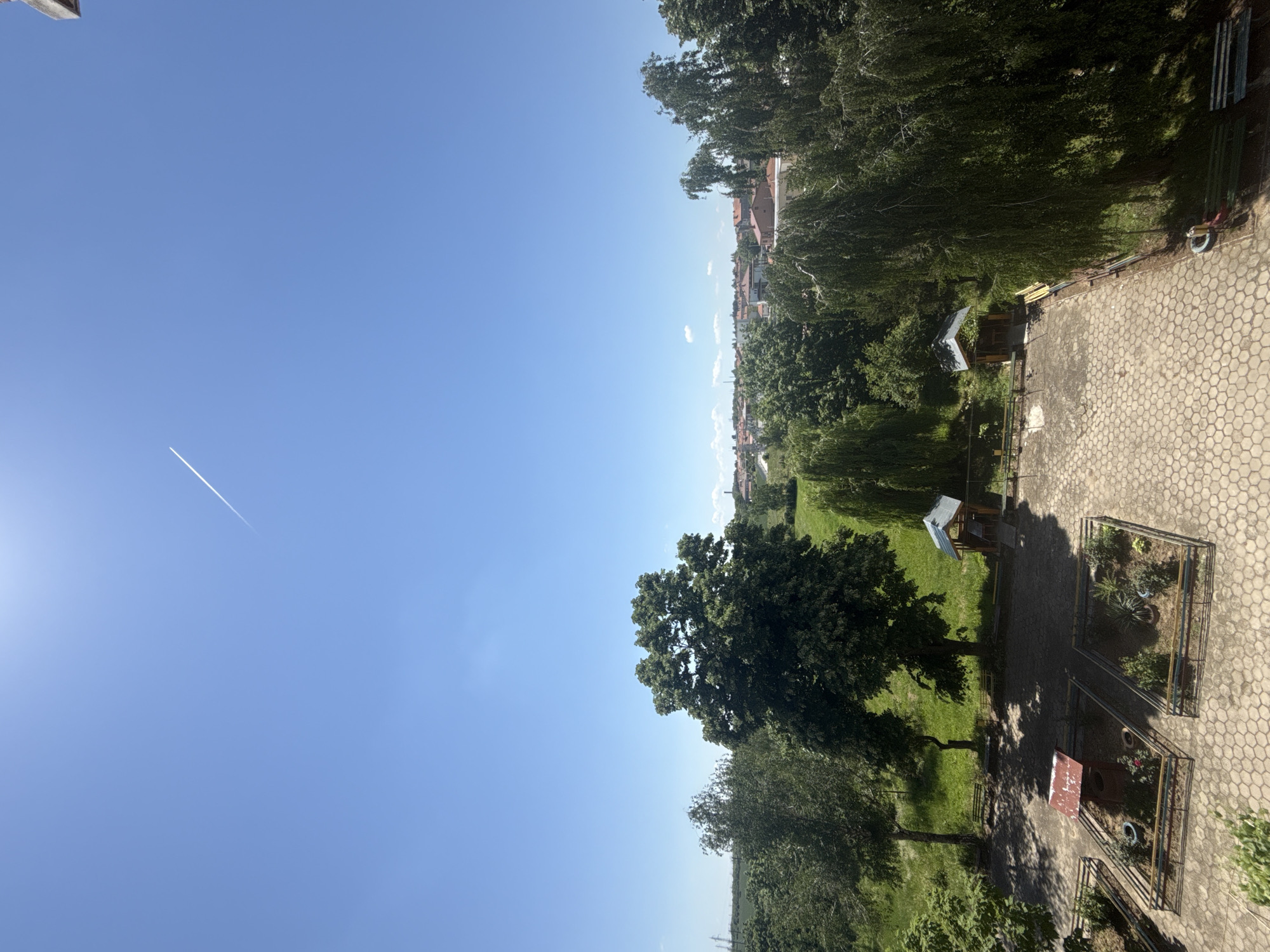 The image depicts a park-like area with trees, green space, and some structures reminiscent of small sheds or playhouses, viewed from a slightly elevated perspective. There are paved areas with a honeycomb pattern, suggesting a walkway or courtyard. The sky is clear blue with a contrail visible. Distant buildings, potentially located in the city of Dulovo, Bulgaria, can be discerned in the background. No people are visible. The general environment appears to be a residential or public green space.