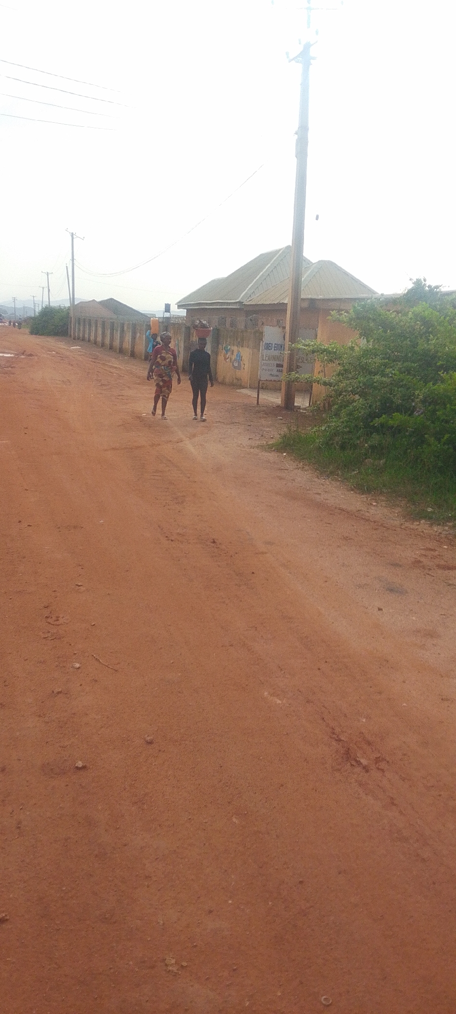 This image depicts a dirt road in the city of Kwang, Nigeria, under a bright, overcast sky. Two individuals are walking on the road. The person on the left, seemingly a woman, wears a patterned dress and carries a large, unseen object balanced on their head. The person on the right, appearing to be a young man, wears a dark, form-fitting outfit and also carries an unseen object on their head.
Flanking the road are buildings and fences. On the right side, a building with a light-colored facade and a metal roof is visible, with a sign partially obscured by foliage. The sign appears to advertise "LOREN-EDOM LEARNING CENTERS". Adjacent to this building is a patch of green grass and bushes. On the left side of the road, a long, beige concrete fence stretches into the distance, with more buildings and what appear to be utility poles and power lines further back. The ground itself is composed of reddish-brown dirt with scattered small stones. The overall environment suggests a rural or semi-urban setting in Nigeria.
