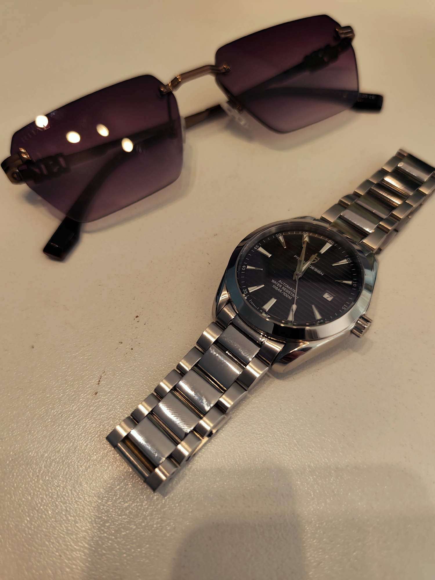 The scene, set in the city of Pazardzhik, Bulgaria, displays a pair of rimless sunglasses and a wristwatch positioned on a light beige, subtly textured surface. The sunglasses are oriented with their left lens prominent, featuring a deep purple tint and reflecting multiple distinct light sources. The frame is minimal, appearing gold-toned, with visible details on the temples. Located to the lower right of the sunglasses, the silver-toned wristwatch, identified as a PAGANI DESIGN, is shown with its metal bracelet unfurled. Its dark, horizontally striped dial is visible, displaying "AUTOMATIC," "WATER RESISTANT," and "10BAR/100M" text, along with luminescent hour markers and hands. A date window is present at the 3 o'clock position. The polished watch case and bracelet links reflect ambient light. The surface exhibits minor dark blemishes and dust particles, particularly noticeable between the two objects.