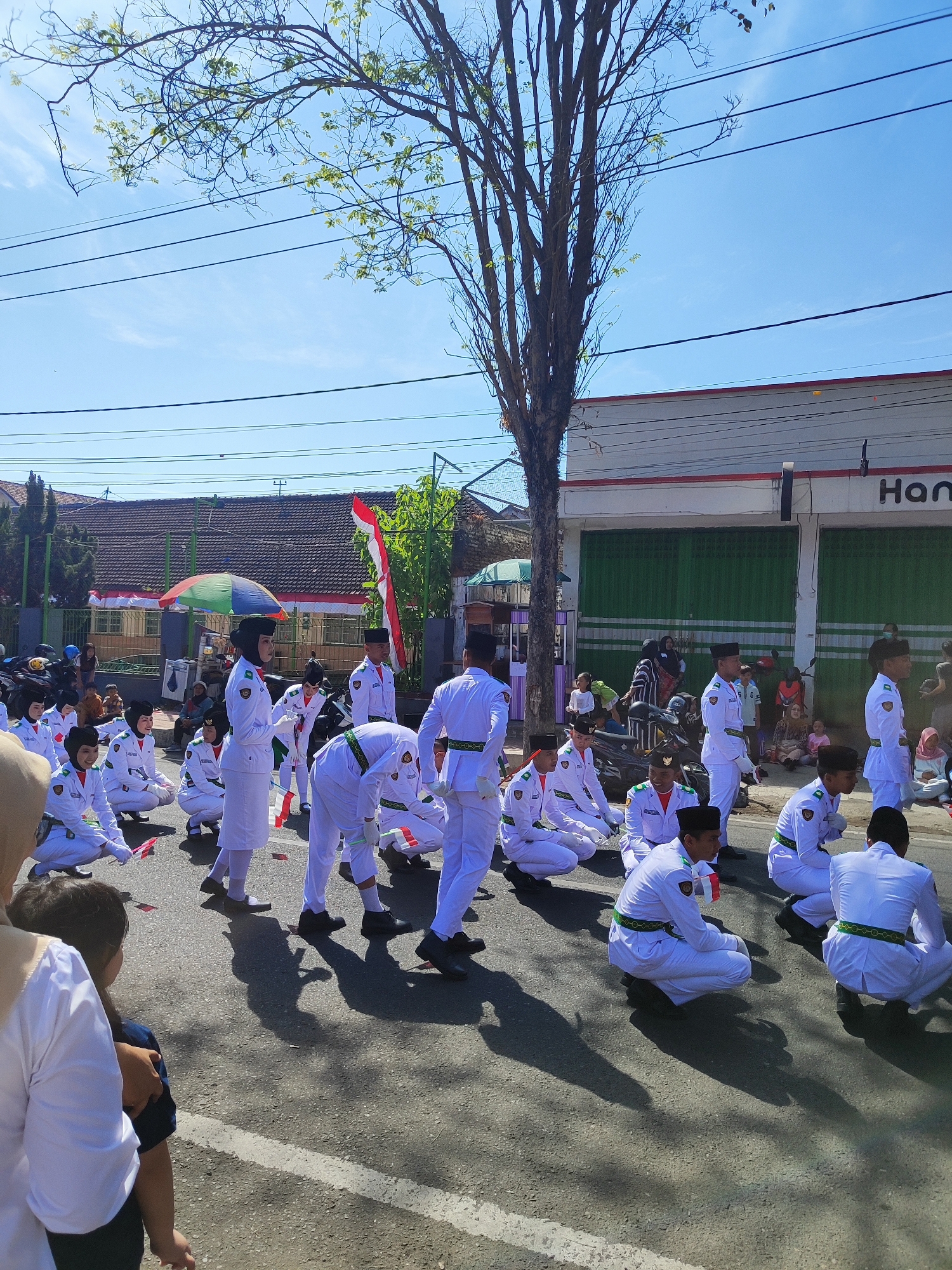 In Dabasah, Indonesia, a group of students dressed in white uniforms with green sashes are participating in a parade. The students, both male and female, are in various stages of a kneeling or squatting position on an asphalt road. Many of them are holding small Indonesian flags. Behind them, spectators are visible, some seated and others standing. The scene is set against a backdrop of buildings, some with green shutters, and a large tree with sparse leaves. Overhead, power lines crisscross the bright blue sky. The atmosphere appears to be one of a celebratory event, likely a national holiday or school event.