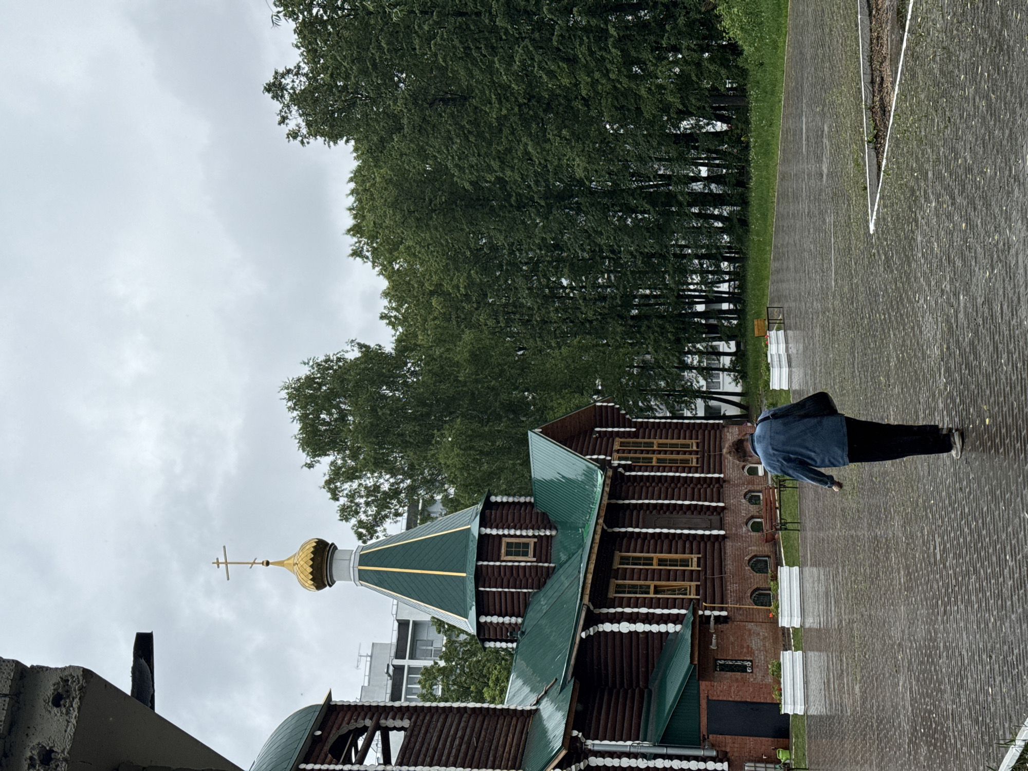 A person in a blue jacket and black pants is walking on a brick sidewalk next to a wooden Russian Orthodox church with green roofs in Chaikovsky, Russia. The church has a golden dome with a cross on top. Behind the church are trees and a cloudy sky. A building can be seen in the background.