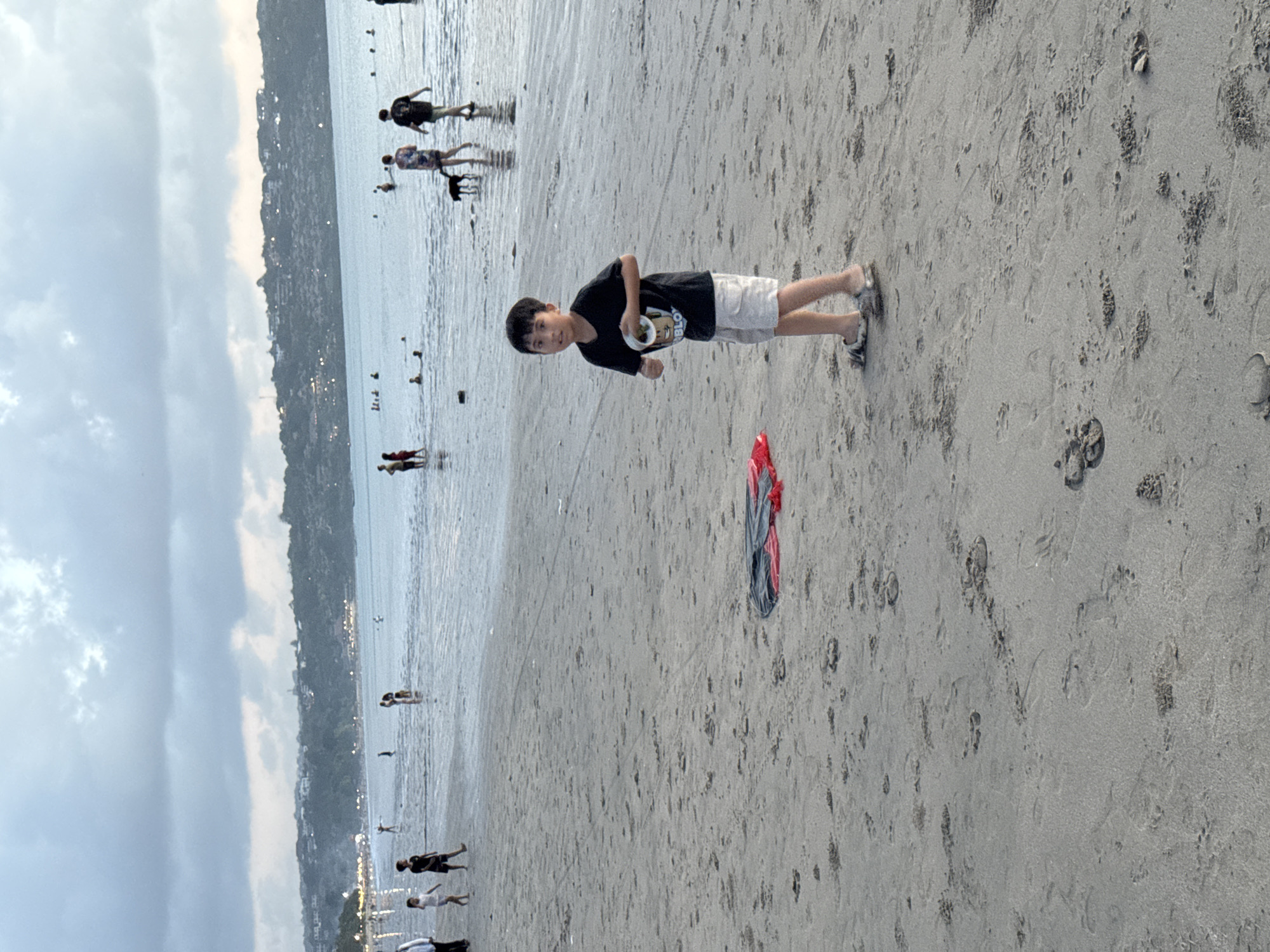 The photograph depicts a beach scene in Kuta Selatan, Indonesia. A young boy stands in the foreground, holding a small, round object in his hand. He is wearing a black t-shirt and light-colored shorts. Behind him, a red and grey garment lies on the sand. The beach is populated with other individuals, some walking along the shoreline and others wading in the water. In the background, a coastline is visible under a cloudy sky.