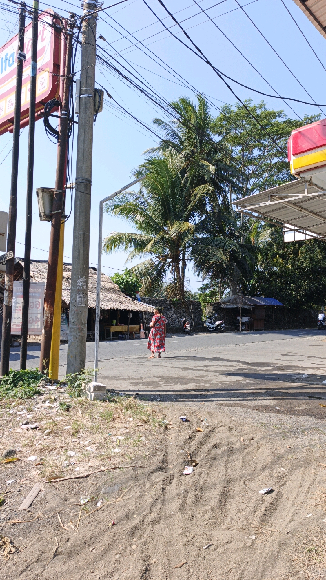 The image depicts a street scene in Desa Batukaras, Indonesia. A woman in a red patterned dress stands on the right side of a paved road, facing left. She is positioned between a tall utility pole with numerous cables and a small, open-fronted building with a thatched roof. Behind her, a palm tree with large fronds towers over the landscape. Further in the background, several small structures with thatched roofs and an open canopy structure are visible. Two motorcycles are parked near one of the buildings. The ground in the foreground is a mix of dirt and sparse vegetation, with some scattered debris. The sky is clear and blue. The overall impression is a rural or semi-rural roadside environment.