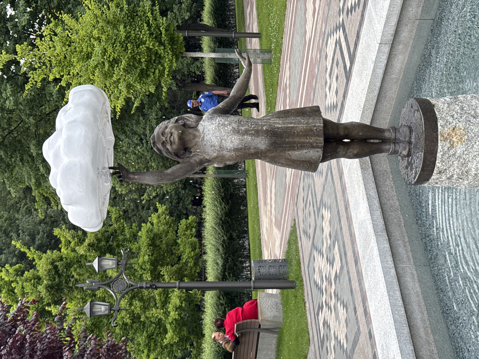 The image shows a bronze statue of a girl reaching up to touch a cloud-shaped fountain element. The statue is located on the edge of a fountain in a park. Behind the statue, there are trees, a park bench with people seated on it, and a lamppost. A person in a blue shirt is walking in the background. The statue and fountain are part of a sculpture called "Hands of the Cloud" by David Yerevantsi, located in the 2750th Anniversary Park in Yerevan, Armenia.