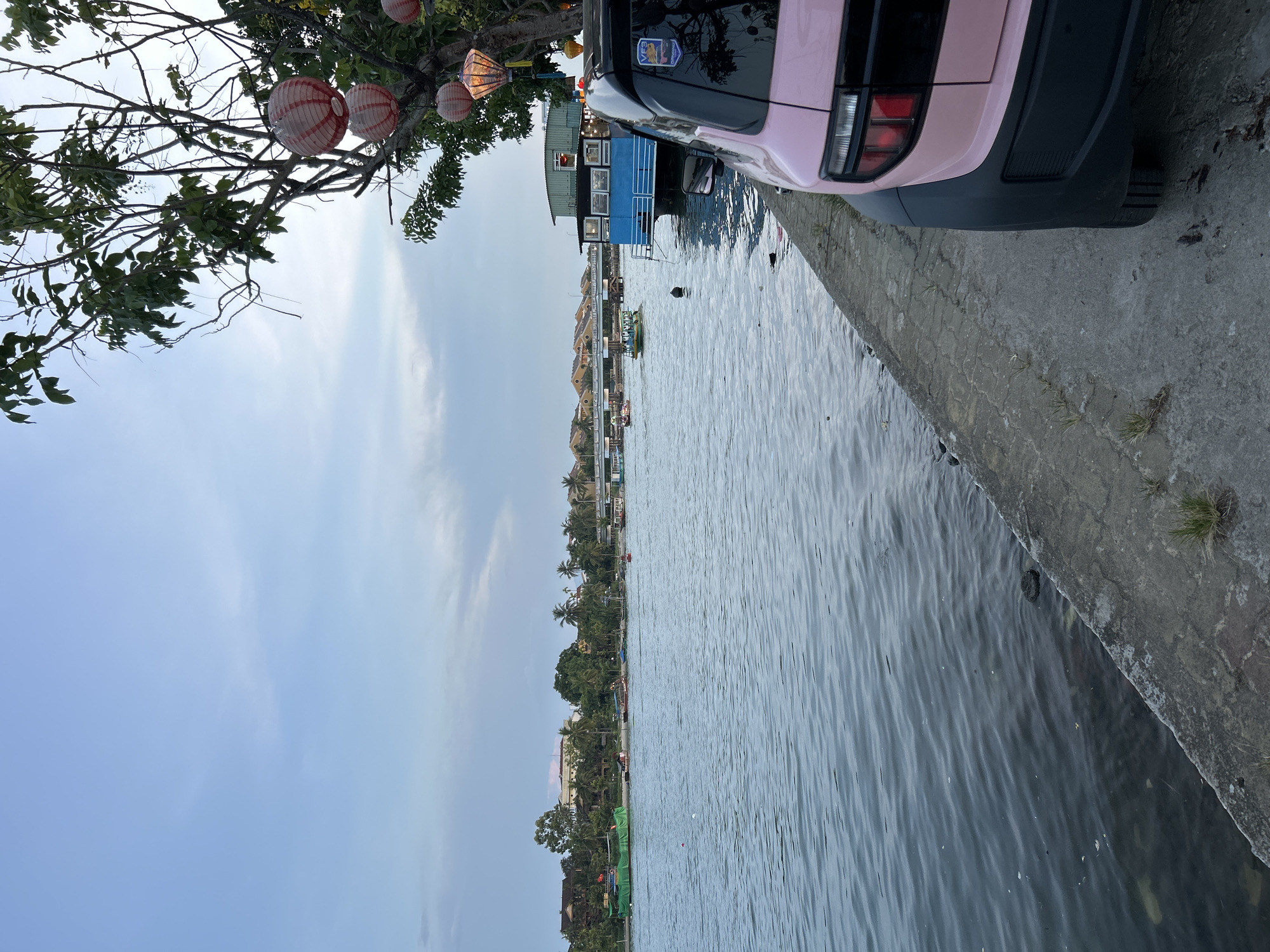 A light pink vehicle is parked on a concrete embankment next to a river. The vehicle's rear is visible, featuring distinctive taillights and black fender flares. The river flows calmly, with several boats visible on its surface. In the background, the riverbank is lined with trees and buildings, including a blue floating structure. Hanging lanterns adorn a tree in the foreground, casting a warm glow. The sky is overcast with soft clouds, suggesting either early morning or late afternoon light. The scene appears to be in Hội An Ward, Vietnam, a region known for its canals and historical architecture.