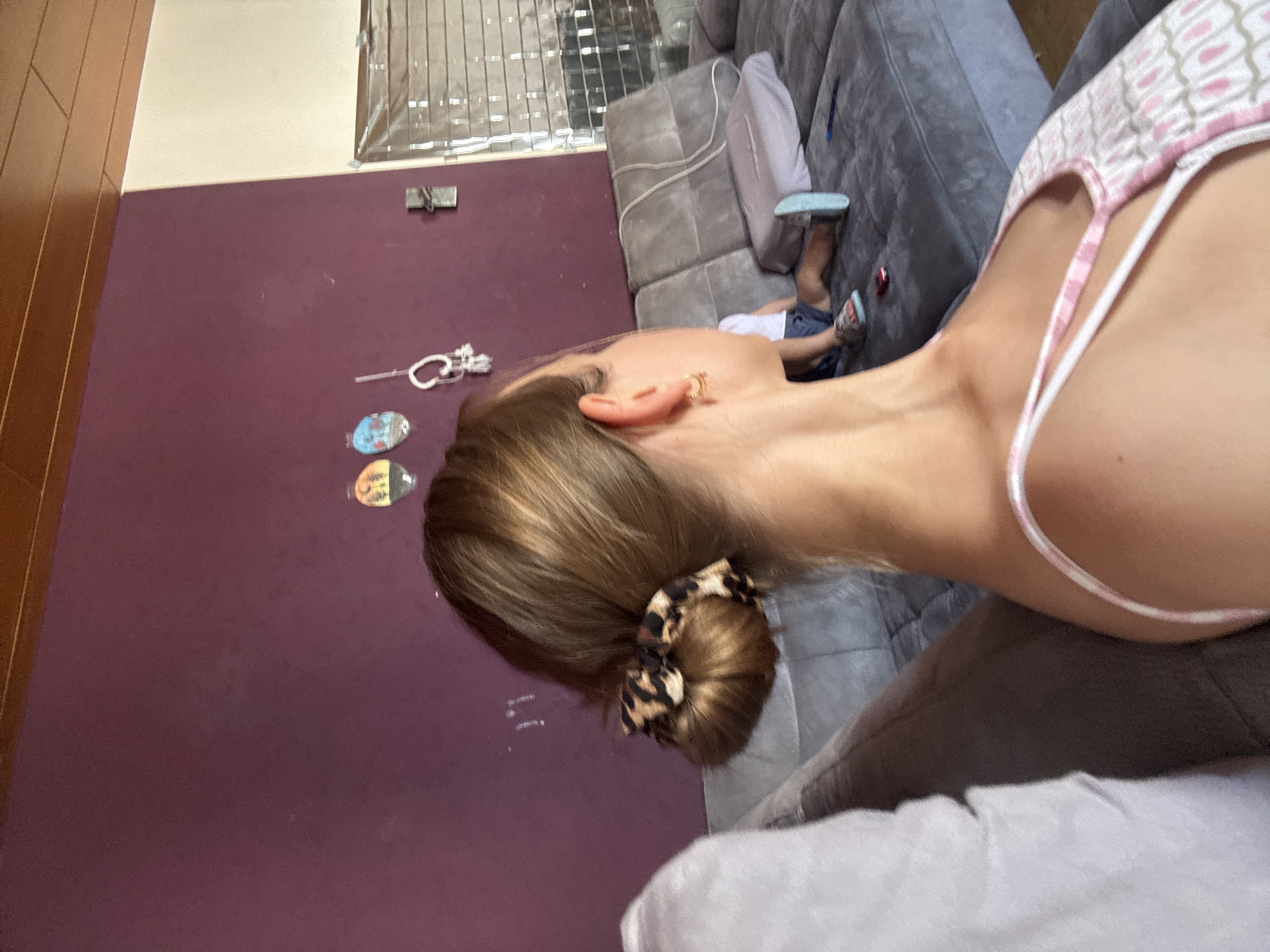 A young woman with light brown hair styled in a bun with a leopard-print scrunchie is shown from the back, seated on a gray couch. She is wearing a white tank top with pink and gray patterns. A child, wearing blue shorts and sandals, is partially visible on the same couch in the background, lying down with their legs extended. The environment appears to be a living room with a purple wall, a wooden ceiling, and a large gray sectional sofa. A window with blinds is visible on the right side of the image. The image was taken in Überherrn, Germany.