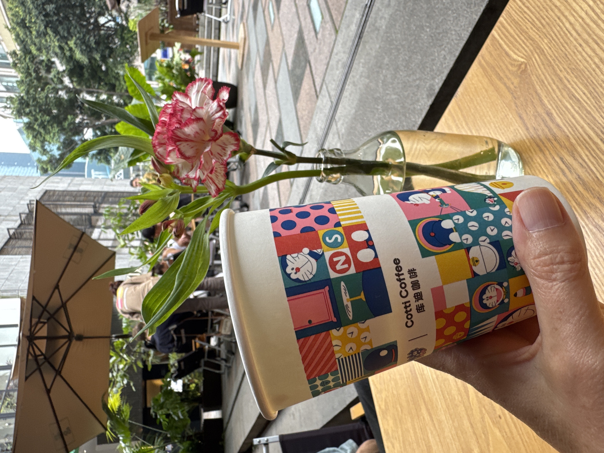 A person holds a paper cup of "Cotti Coffee" beside a small glass vase with a pink and white carnation. The scene is likely an outdoor seating area, as visible in the background are other people sitting under umbrellas, trees, and surrounding buildings. The location might be Yuzhong District, China, based on the "Cotti Coffee" brand's presence in the area. The cup has a cartoon design on it. The table is a light brown wood.