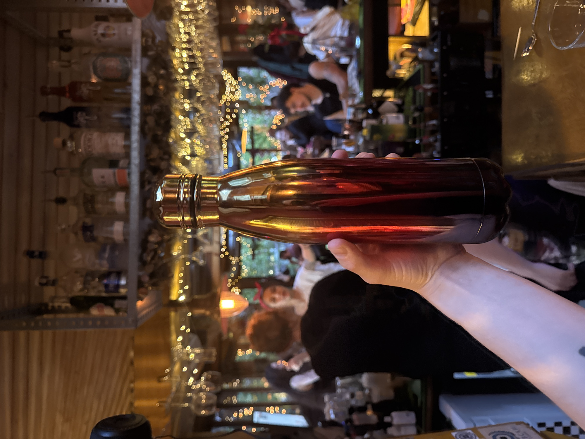 The image displays an indoor bar scene, rotated 90 degrees clockwise. In the foreground, a person's light-skinned hand, bearing a faint tattoo on the wrist, firmly grips a shiny, reddish-brown metallic bottle or thermos horizontally. The bottle reflects the warm ambient lighting of the establishment.
To the left of the hand, a metal shelf unit is visible, holding several liquor bottles. One bottle on the top shelf is clearly identifiable as "Malibu." Other bottles are too blurry to read their labels.
The background is heavily blurred, creating a bokeh effect from numerous warm, circular lights, likely decorative string lights. Behind the prominent bottle and the lights, two individuals are discernible. One person appears to be a bartender or staff member, leaning forward behind a bar counter. Further back, another person with what looks like red hair or a red hair accessory is vaguely visible.
The setting is a dimly lit bar or pub, characterized by a wooden slatted wall on the far left and various bar equipment and glasses in the blurred background. The warm, artificial lighting suggests it is evening or night. No specific activities are clearly defined beyond holding the bottle, but the context implies socializing or working within a bar. The location context is Sofia, Bulgaria.
