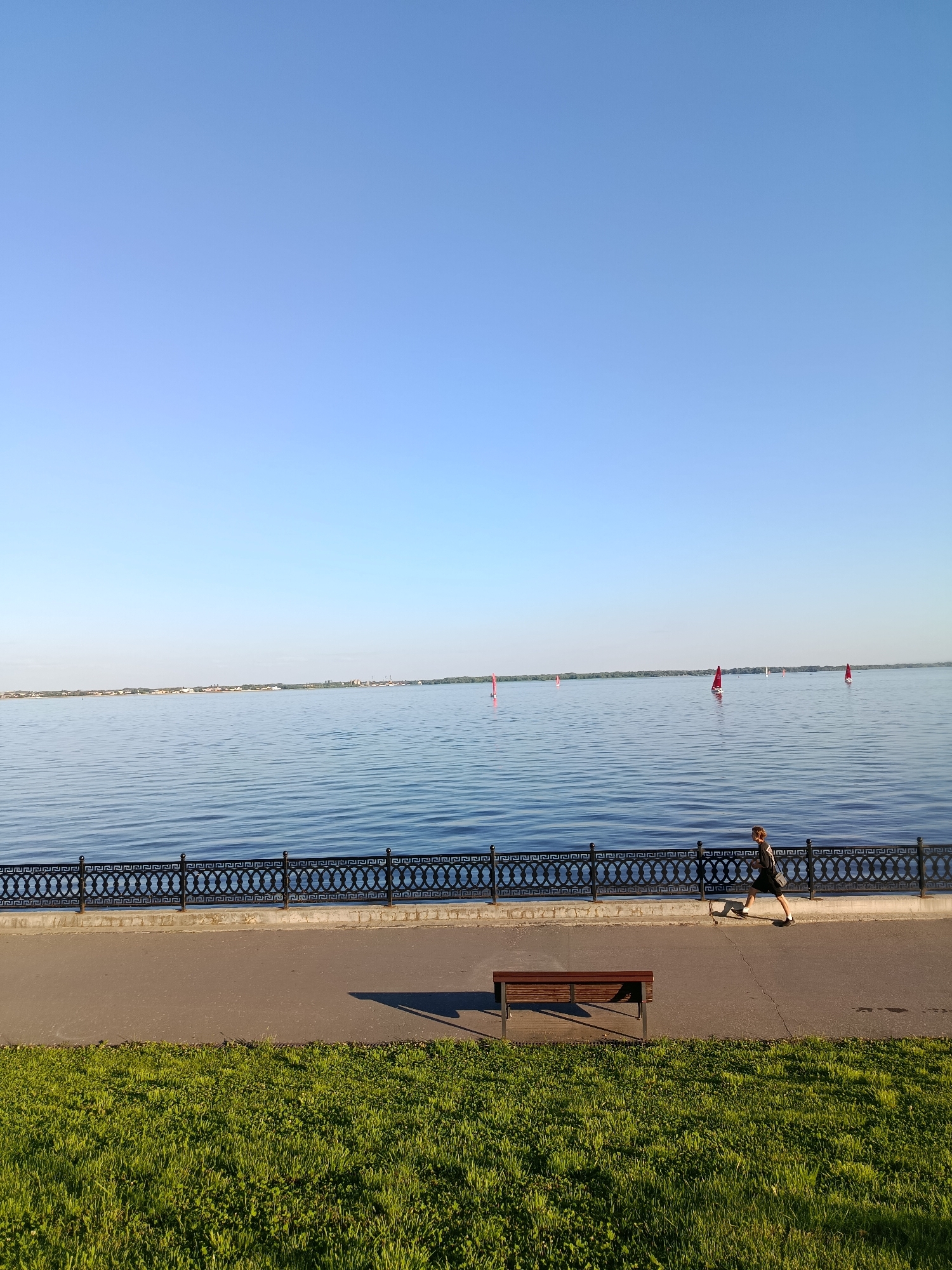 A person is jogging along a waterfront promenade next to a wide, calm body of water under a clear blue sky. The scene suggests a sunny day, likely in the morning or late afternoon due to the long shadows cast by the bench and the runner. The runner, seen in profile, is wearing dark athletic clothing and appears to be mid-stride.
The promenade is bordered by a decorative black metal fence with an ornate geometric pattern. Beyond the fence, the water stretches to the horizon, where a distant tree line and some buildings are visible. Several small sailboats with red sails are scattered across the water, adding a touch of color and activity to the scene. A wooden bench sits empty on the promenade, its shadow stretching across the asphalt. In the foreground, a strip of vibrant green grass completes the image. The overall atmosphere is peaceful and serene, typical of an early morning or evening outdoor scene in a park or along a riverbank. No visible text is present in the image. The location context of Saratov, Russia, implies this is likely a scene along the Volga River.