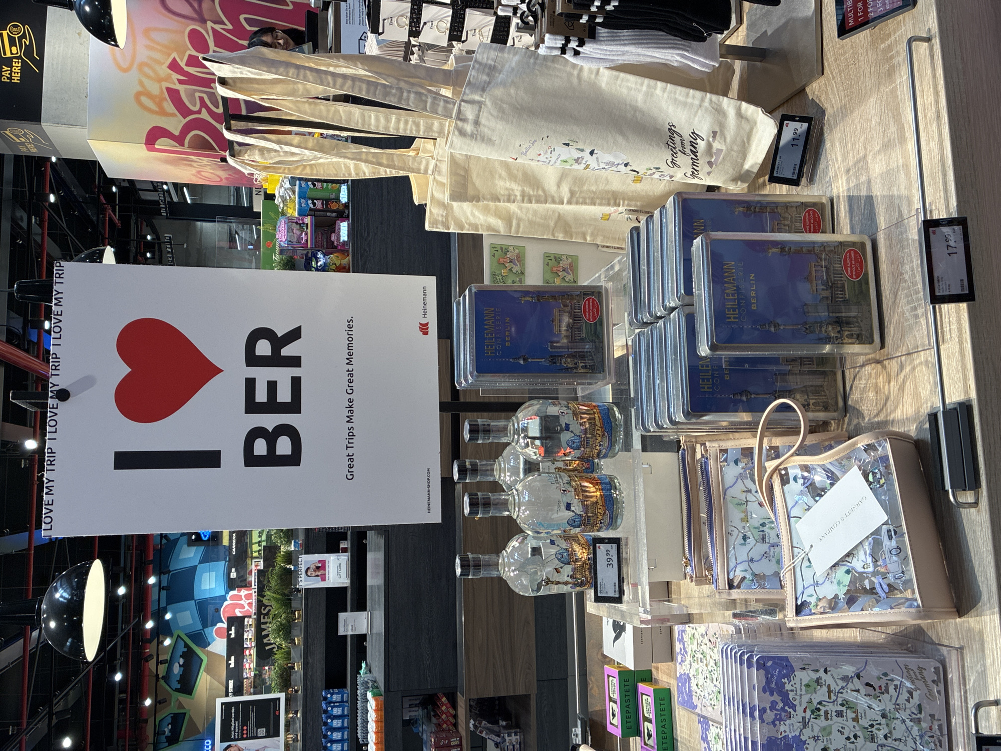 The image shows a retail display area inside a duty-free shop at Berlin Brandenburg Airport, Schönefeld, Germany. A large white sign with the text "I BER" and a red heart is prominently displayed, with the slogan "Great Trips Make Great Memories." Below the sign, several items are arranged on a wooden counter. These include four bottles of clear liquid, possibly spirits, each with a label depicting a city scene. To the right, three canvas tote bags with "Greetings from Germany" printed on them are displayed. In front of the tote bags are several small, rectangular boxes labeled "HEILEMANN CONFISERIE BERLIN." There are also several transparent pouches with a map design. Further to the left, there are various small packaged items, including what appear to be candy boxes, and a stack of notebooks or maps with a city map design. A price tag of €39.99 is visible near the bottles. A price tag of €11.99 is visible near the "HEILEMANN" boxes. Another price tag of €17.99 is visible on the lower part of the counter. In the background, shelves are stocked with various products, and overhead lighting illuminates the scene. A person with glasses is partially visible in the background, near the tote bags.