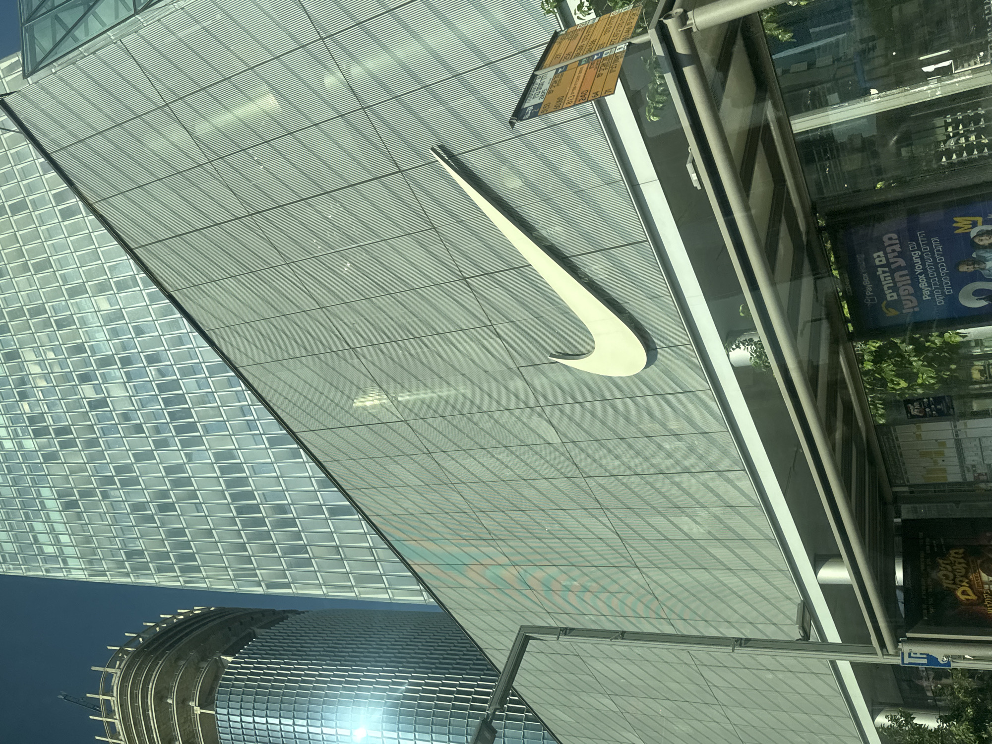 The image depicts a street-level view of the Azrieli Center towers in Tel Aviv, Israel, dominated by a large Nike swoosh logo on the facade of one of the buildings. The perspective is from a bus stop, with the roof and seating area of the bus stop partially obscuring the lower portion of the Nike building and the street.
The Azrieli Center complex is characterized by its modern architecture, featuring glass and metal facades. The tower on the left appears to be under construction or renovation, with scaffolding visible at its upper levels. The central tower displays a grid-like pattern of windows, reflecting the bright blue sky and sunlight. The Nike logo, prominently displayed in white, spans a significant portion of the visible facade of the Nike-branded building.
Adjacent to the Nike building, at the street level and integrated with the bus stop structure, is a series of advertisements. One prominent advertisement, visible through the glass of the bus stop, is in Hebrew and appears to be for "PayBox Young." This suggests the location is a commercial area with active advertising.
The environment is urban, with clear daylight conditions. The presence of a bus stop indicates a public transportation hub within the city. The image captures a slice of urban life in Tel Aviv, highlighting iconic architecture and commercial branding.