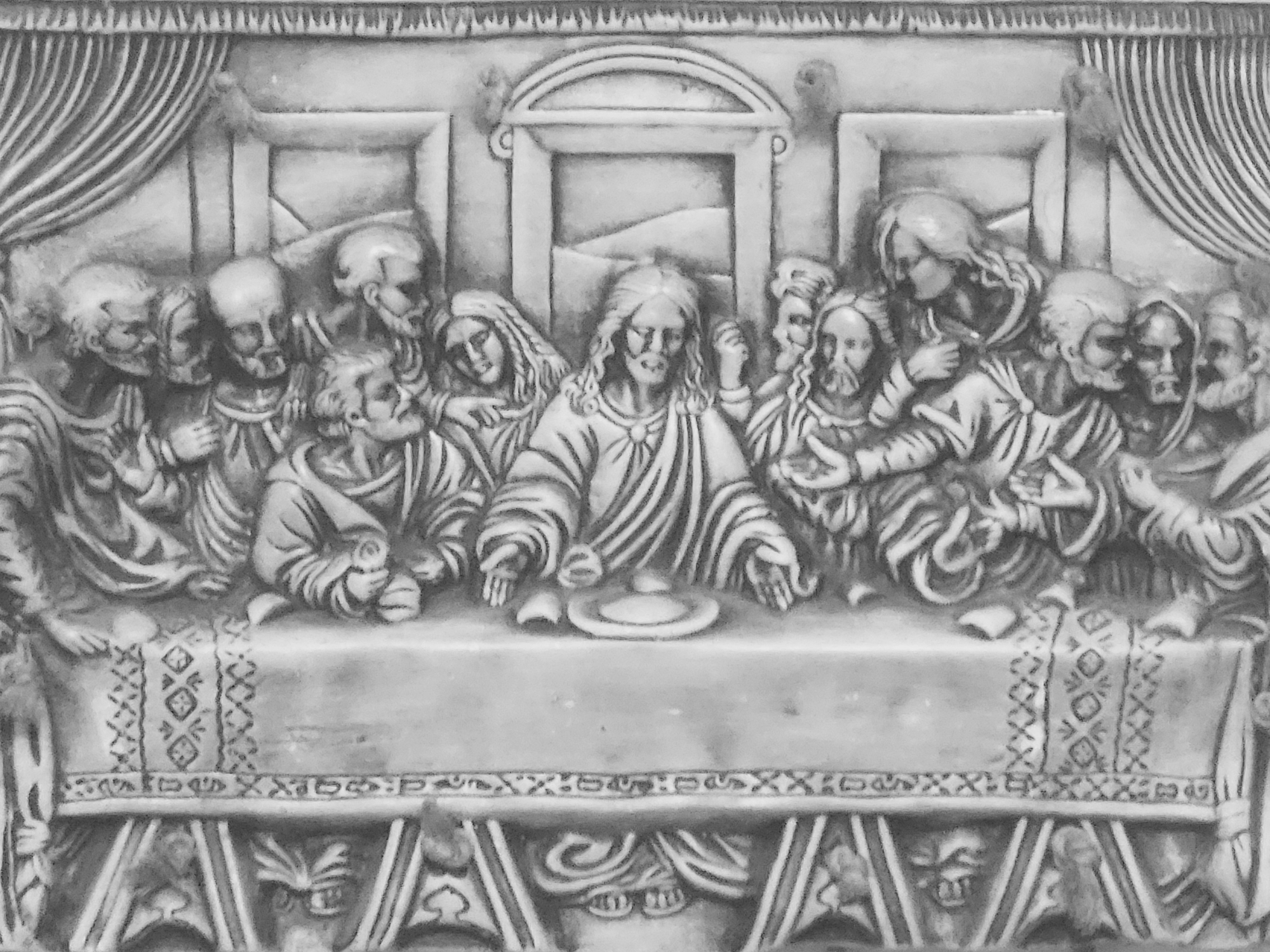 The image is a grayscale depiction of "The Last Supper," likely a sculpture or relief carving. Thirteen figures are seated at a long table, the central figure being Jesus. He has long hair and is wearing a robe, with hands outstretched over a plate and goblet. The other figures are arranged on either side of him, also wearing robes, and appear to be in conversation or reacting to Jesus.
The table is draped with a cloth decorated with geometric patterns. The base of the table is adorned with architectural details and carved motifs.
The background includes draped curtains on the sides and framed panels above the figures' heads. The central panel has a decorative arch.
It is not possible to determine the location in Yerevan, Armenia from the image.
