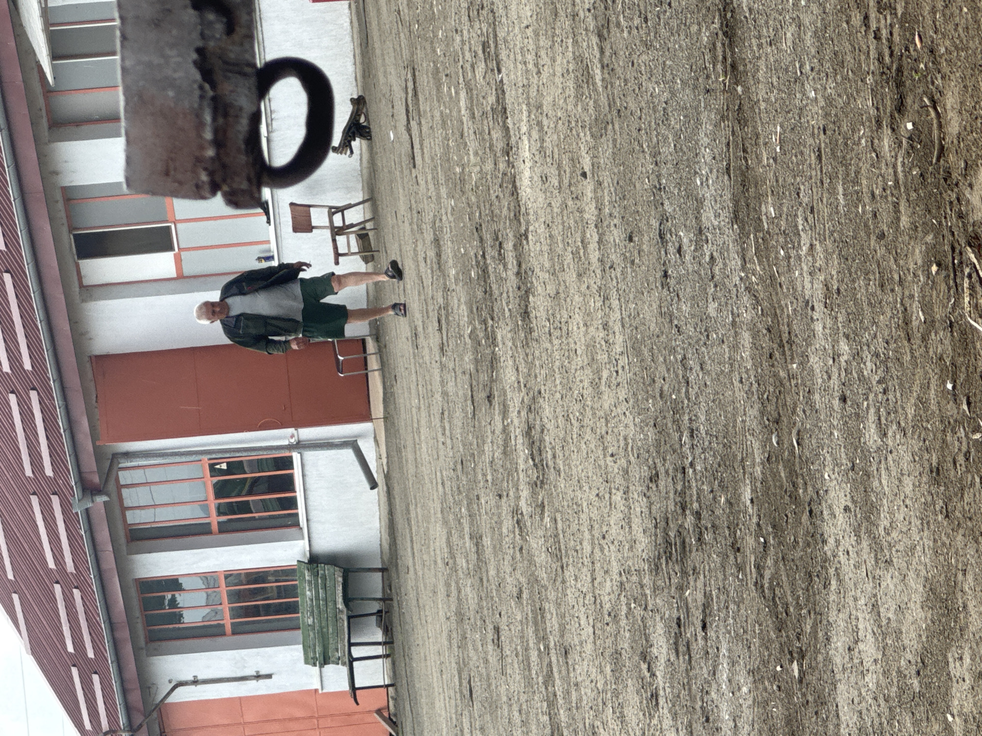 A man is walking in the open space between a building and a gravel area, possibly in Sarnevo, Bulgaria. The building has red roofs, red doors, and windows with red trim. Various chairs and a small table are visible. A black, circular object is hanging in the upper left corner of the image.
