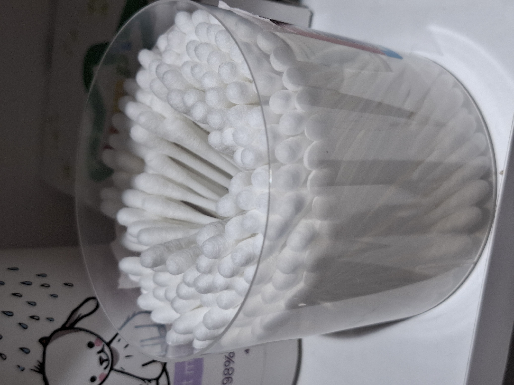 The image displays a clear, cylindrical plastic container filled with numerous white cotton swabs (Q-tips), positioned sideways. The cotton swabs are densely packed, with their cotton-tipped ends facing upwards and outwards, creating a full, fan-like arrangement within the container. The transparency of the container allows for a clear view of the items inside.
In the lower foreground, partially visible, is another item, possibly a baby product, featuring a cartoon illustration of a white bunny with pink cheeks and black outlines. Above the bunny, small blue droplet shapes are visible, resembling raindrops or teardrops. Part of the text "98%" is visible on this item. In the background, beyond the cotton swab container, faint outlines of other objects or packaging can be seen, though they are out of focus and indistinct. The overall setting appears to be indoors. No activity is taking place; it's a still life arrangement of personal care items.