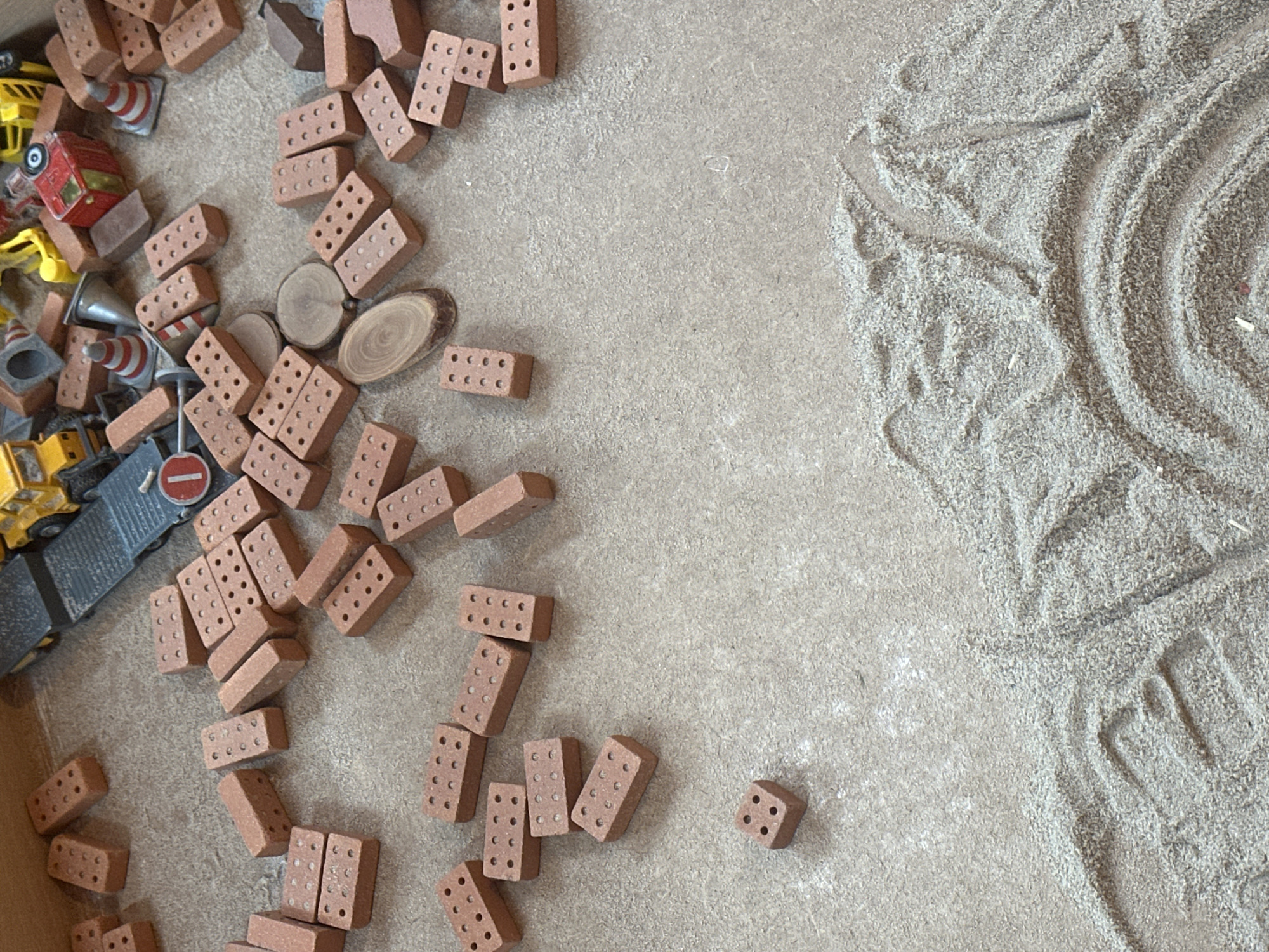 The image shows a collection of miniature construction toys, including red bricks, a yellow dump truck, traffic cones, and other small construction vehicles. These toys are scattered on a flat, textured surface that resembles sand or fine gravel. In the foreground, there are patterns raked into the sand, suggesting a zen garden or a sand play area. The lighting is natural, casting soft shadows. There are no people visible in the image.