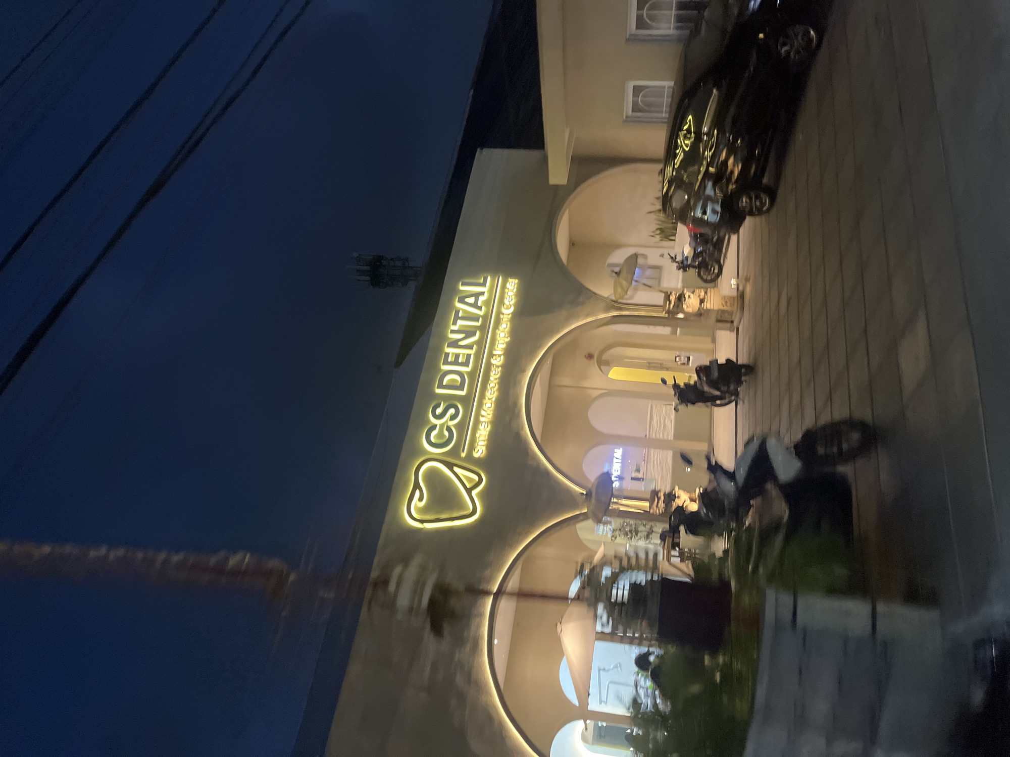 The image shows the exterior of a dental clinic called "CS DENTAL Smile Makeover & Implant Center" in North Kuta, Indonesia. The clinic's facade is illuminated by bright yellow neon lights in the shape of a tooth and the text of the clinic's name. The building has arched entrances and a modern design. Several motorbikes are parked outside the clinic, and a black car is also visible. The scene is captured at dusk or night, with the sky appearing dark blue. The overall impression is of a well-lit, modern establishment in a semi-urban or commercial area.