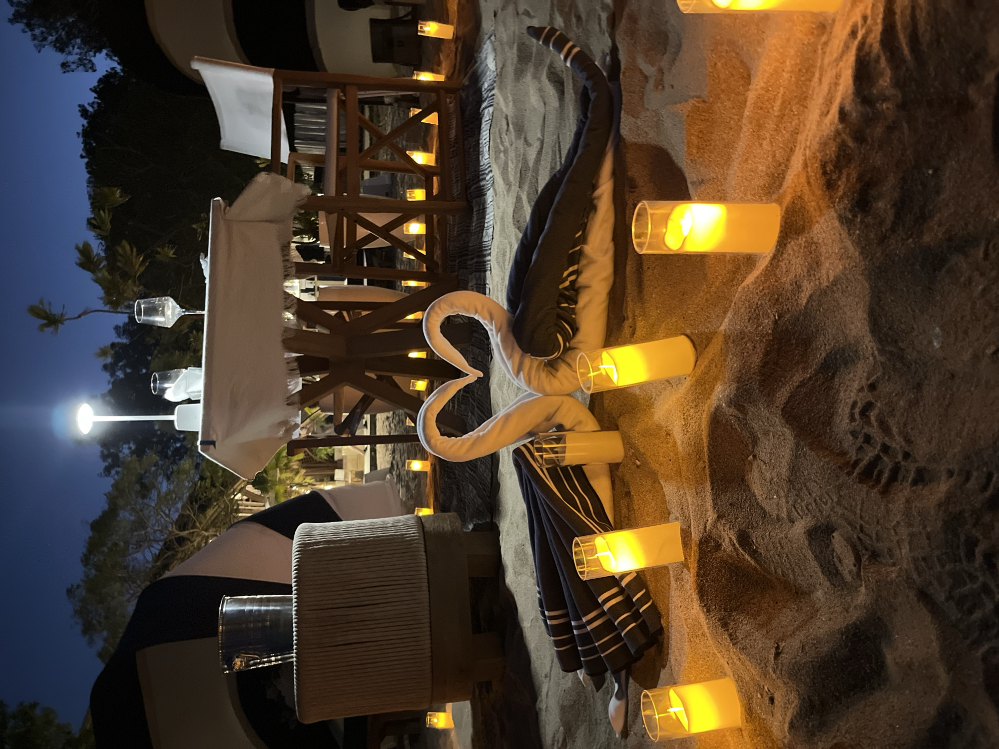 A romantic beach dinner setup at night features a table draped with a white cloth, set for two with wine glasses. In the foreground, two swan-shaped towel art figures face each other, their bodies forming a heart shape. They are adorned with striped fabric and accompanied by two cylindrical glasses with a yellow liquid. Numerous pillar candles in glass holders are strategically placed around the sand, illuminating the scene. In the background, trees and a tent-like structure are visible. The location is the city of Kuta Selatan, Indonesia.