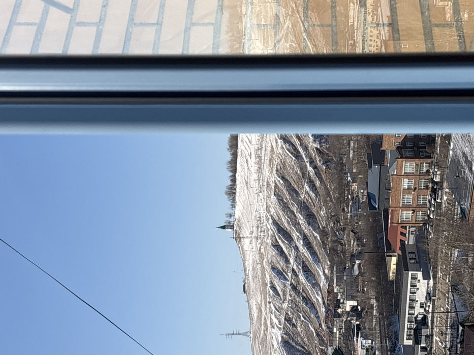 The image displays an elevated view of Krasnoyarsk, Russia, on a clear, sunny day with a blue sky. The scene is partially framed on the right by a vertical light-blue window mullion, with a reflection of a light-brown brick building visible on the adjacent glass surface. A dark, thin cable traverses the upper left quadrant.
In the mid-ground, a sprawling urban area with various buildings is visible. These include multi-story brick structures with multiple windows and visible parked vehicles, as well as darker-roofed commercial or industrial buildings. Patches of snow are present on rooftops and ground surfaces within the urban layout.
The background is dominated by a snow-dusted hill, identified as Karaulnaya Mountain, featuring distinct horizontal snow patterns on its slopes. At the summit stands the Paraskeva Pyatnitsa Chapel, a white building with a green conical roof and spire. To the left of the chapel, on a lower ridge of the hill, a metal lattice power transmission tower is visible. Scattered bare trees populate the hill and urban areas. No individuals or specific actions are depicted within the frame.