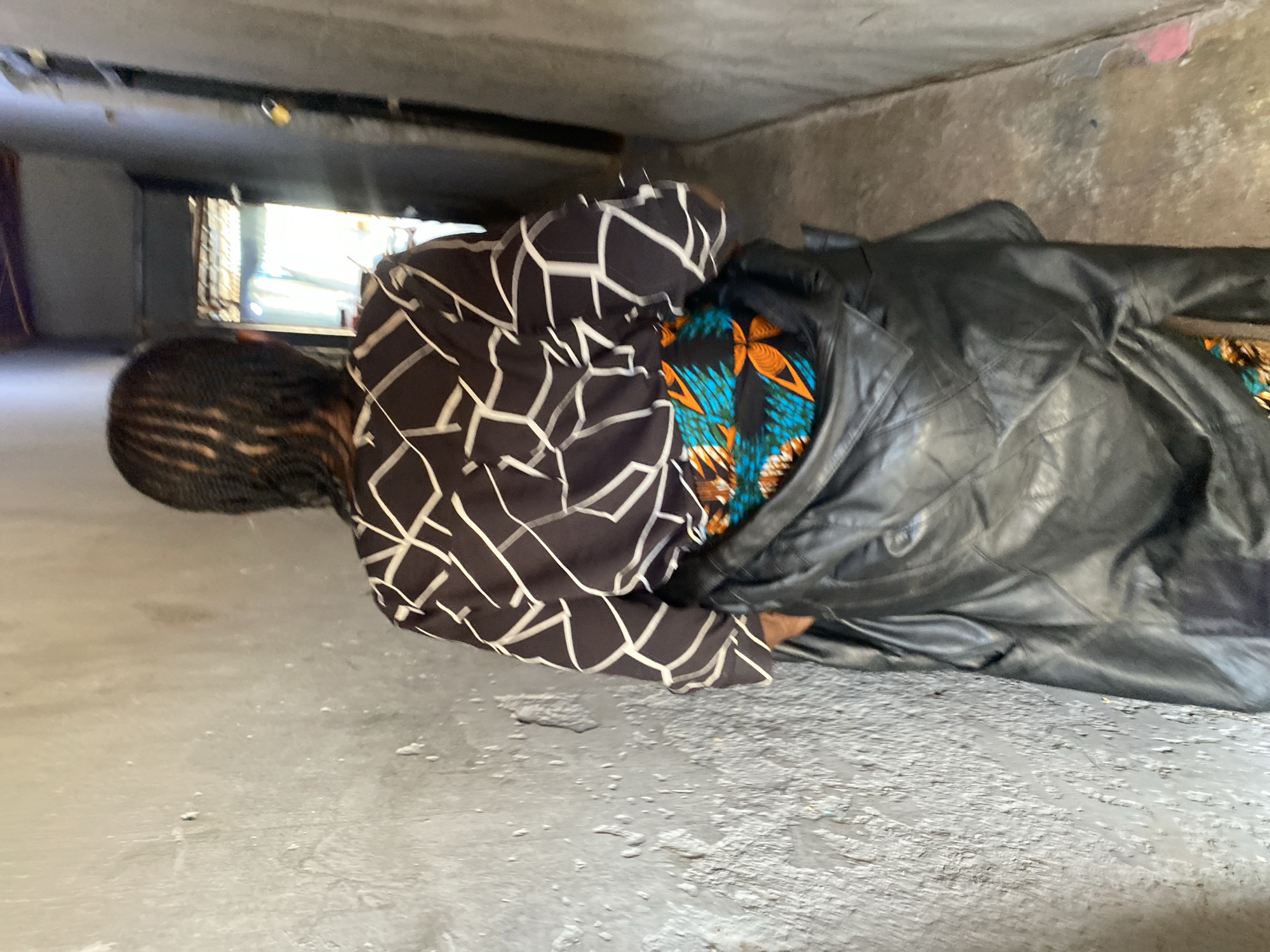 In a narrow hallway with textured, light gray walls, a person seen from behind walks away from the camera. They have braided hair and wear a dark long-sleeved shirt with a white geometric pattern. A black jacket is draped over their shoulders, and a colorful fabric with orange, blue, and black patterns is visible underneath. The hallway opens to a brighter area with corrugated metal roofing and sunlight. The scene appears to be in a residential area in Jos, Nigeria.