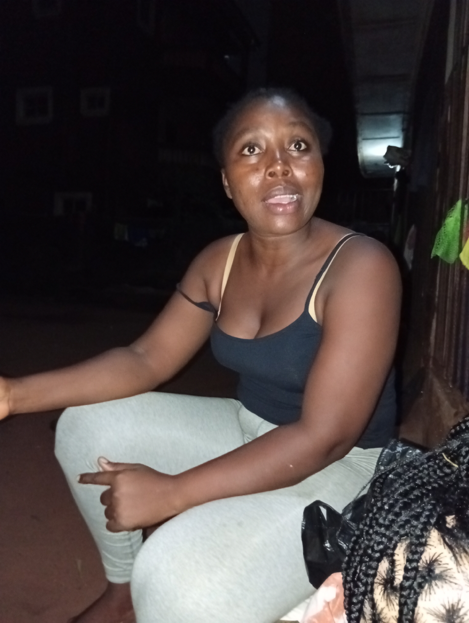 The image shows a woman sitting and gesturing with her right hand. She has dark skin and is wearing a black tank top with light-colored leggings. Her hair is pulled back from her face. A braided wig in a black plastic bag rests beside her. The setting appears to be outdoors at night in the city of Ogidi, Nigeria. Dark buildings are partially visible in the background. There is a fence to her right.