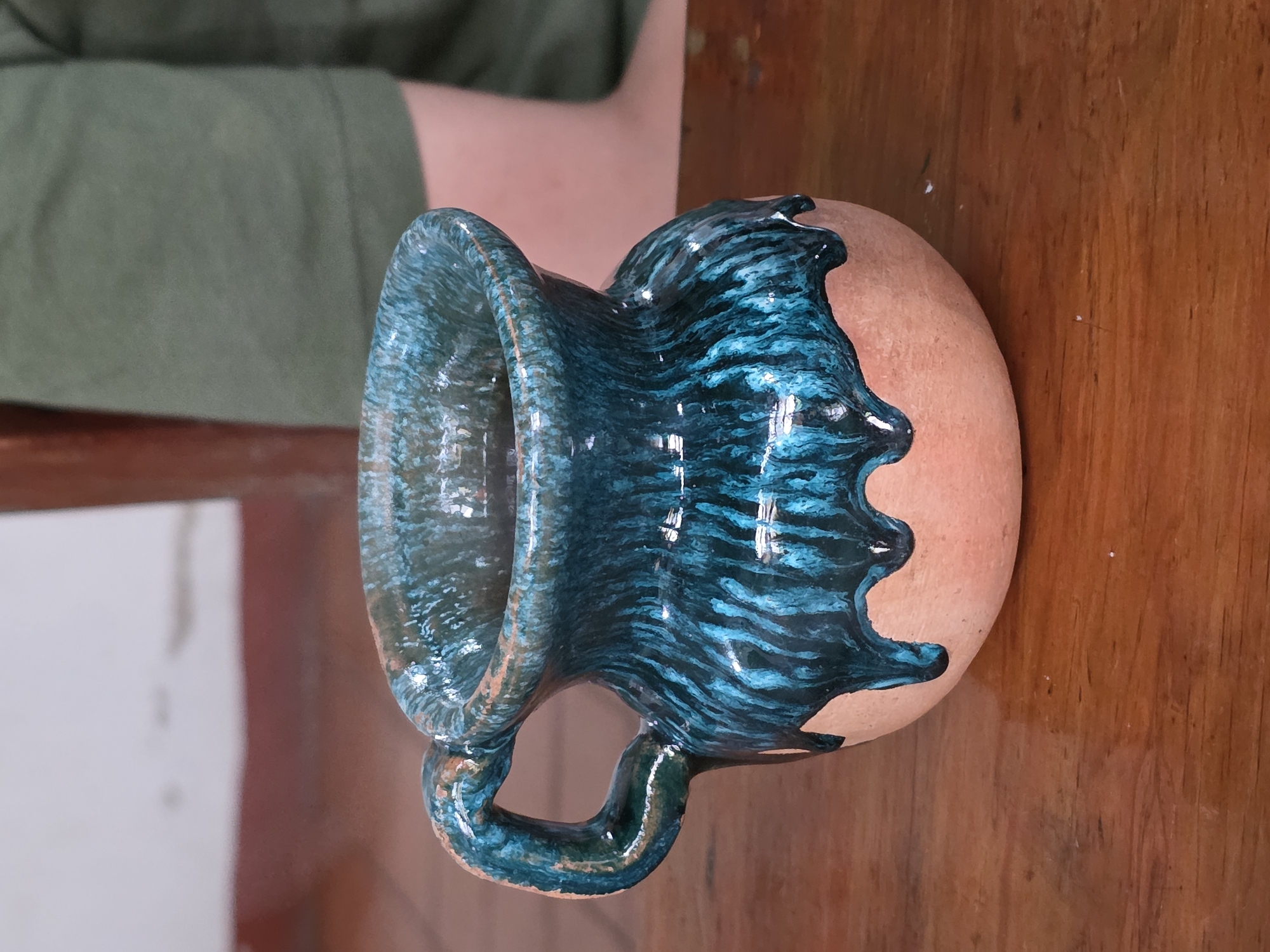 A hand-crafted ceramic mug sits on a wooden surface. The mug is partially glazed, with a blue-green glaze covering the handle, the flared upper section, and dripping down the side of the rounded, unglazed terracotta body. The wood surface has a shiny finish. In the background, a person wearing a green shirt is partially visible. The scene could be located in the city of Cuetzalan, Mexico.