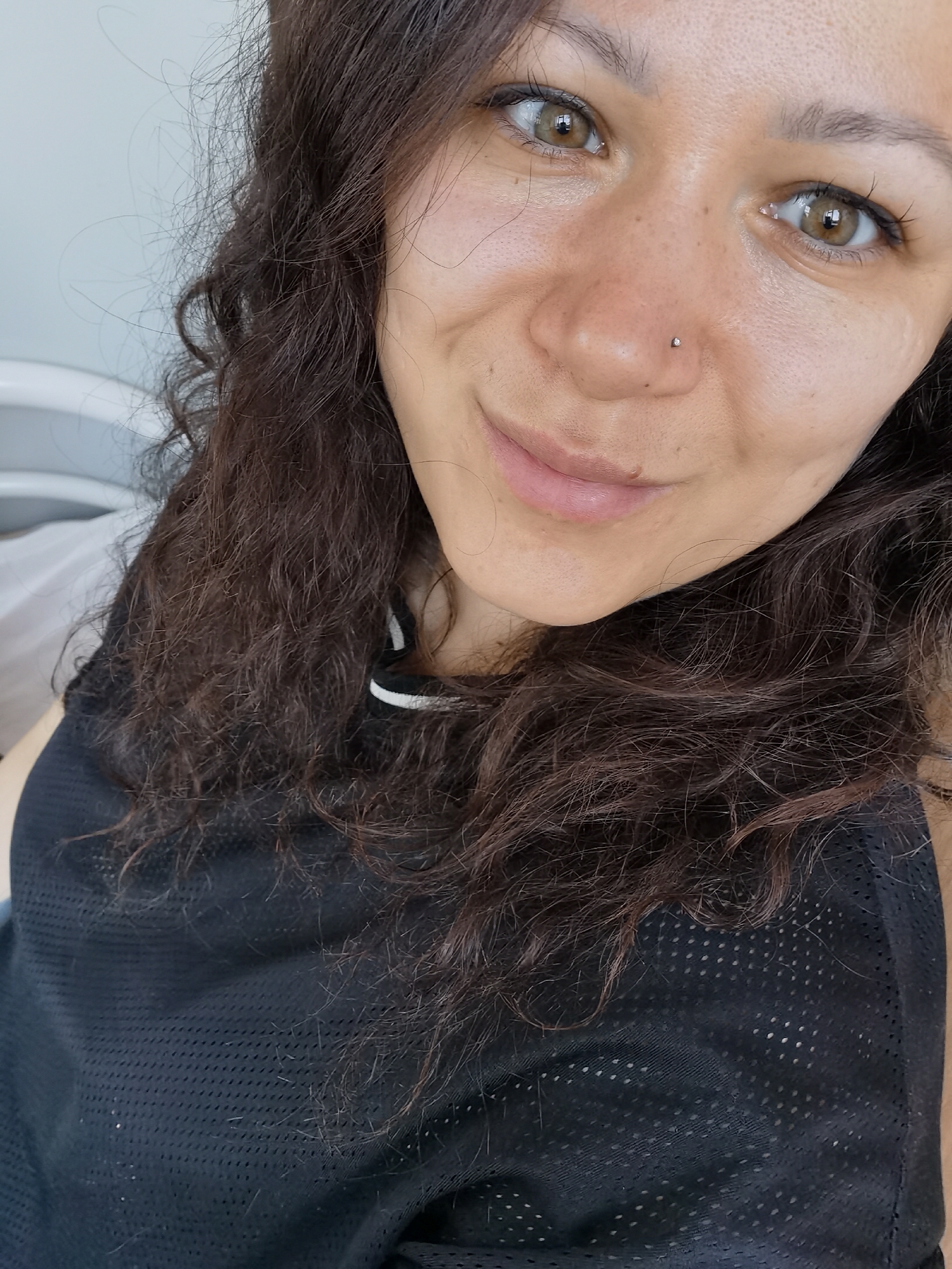 A close-up portrait features a woman with curly brown hair, light brown eyes, and a nose piercing. She is wearing a black, mesh-like tank top with a white trim. The background is indistinct, but appears to be a light-colored wall. The woman's expression suggests a slight smile. There is no indication of the location being in Perm, Russia.