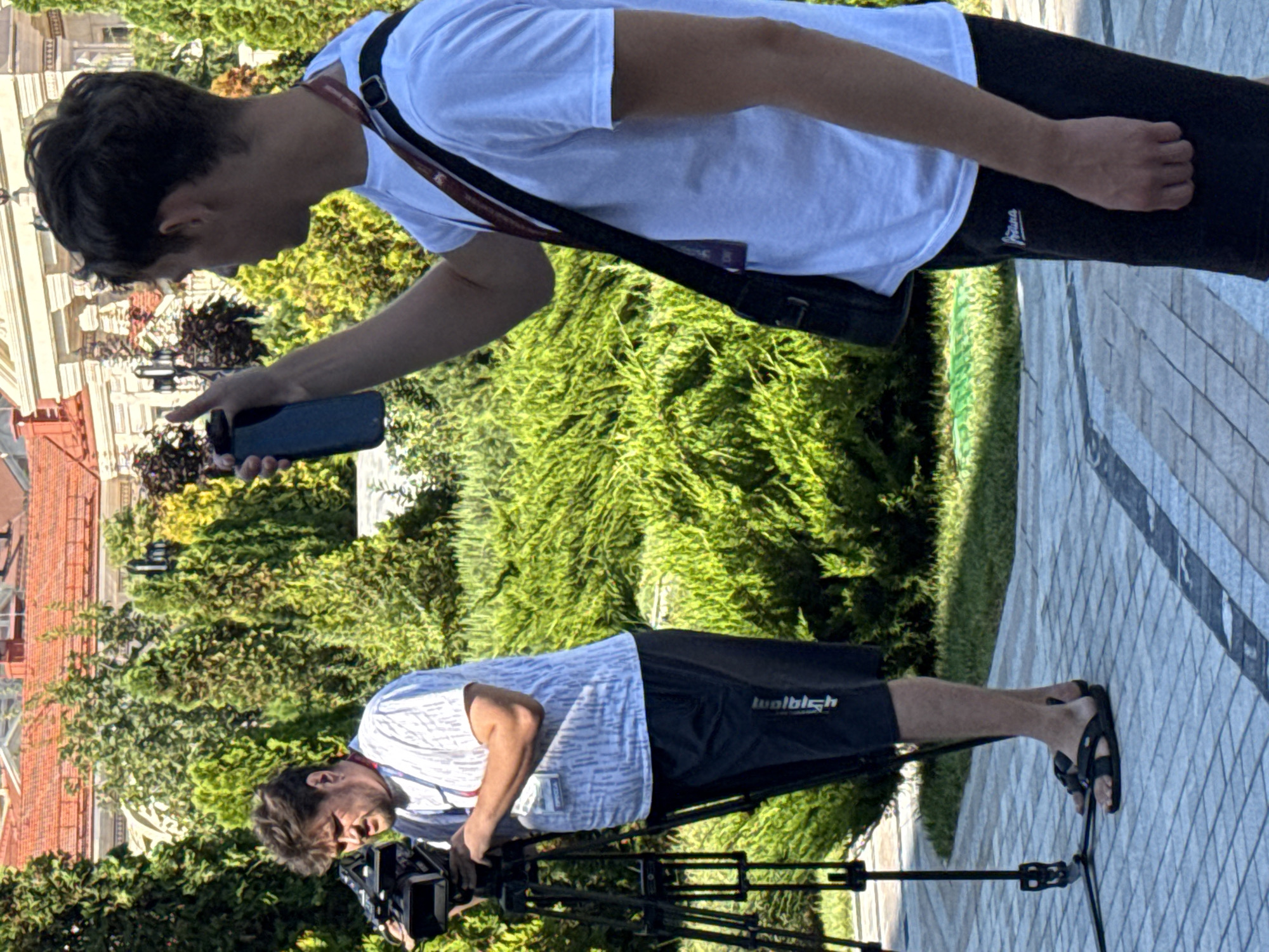 Two men are present in a manicured outdoor setting with green bushes and trees in the background. A building with a red tiled roof and classical architectural elements is visible behind the foliage. The ground is paved with a patterned stone walkway.
The man on the left, positioned behind a professional video camera mounted on a tripod, is wearing a white t-shirt with a blue striped pattern and black shorts. He is looking towards the camera, holding a microphone and a camera controller. He has dark, tousled hair and a beard. He is wearing black flip-flops.
The man on the right, facing away from the camera, is wearing a white t-shirt, black shorts, and a black shoulder bag with a red strap. He is holding a dark-colored water bottle in his right hand, with his arm extended upwards. He has short, dark hair. He appears to be filming or photographing something with his phone, which is held in his left hand.
The overall scene suggests a media event or filming session taking place outdoors in a city park or public space.