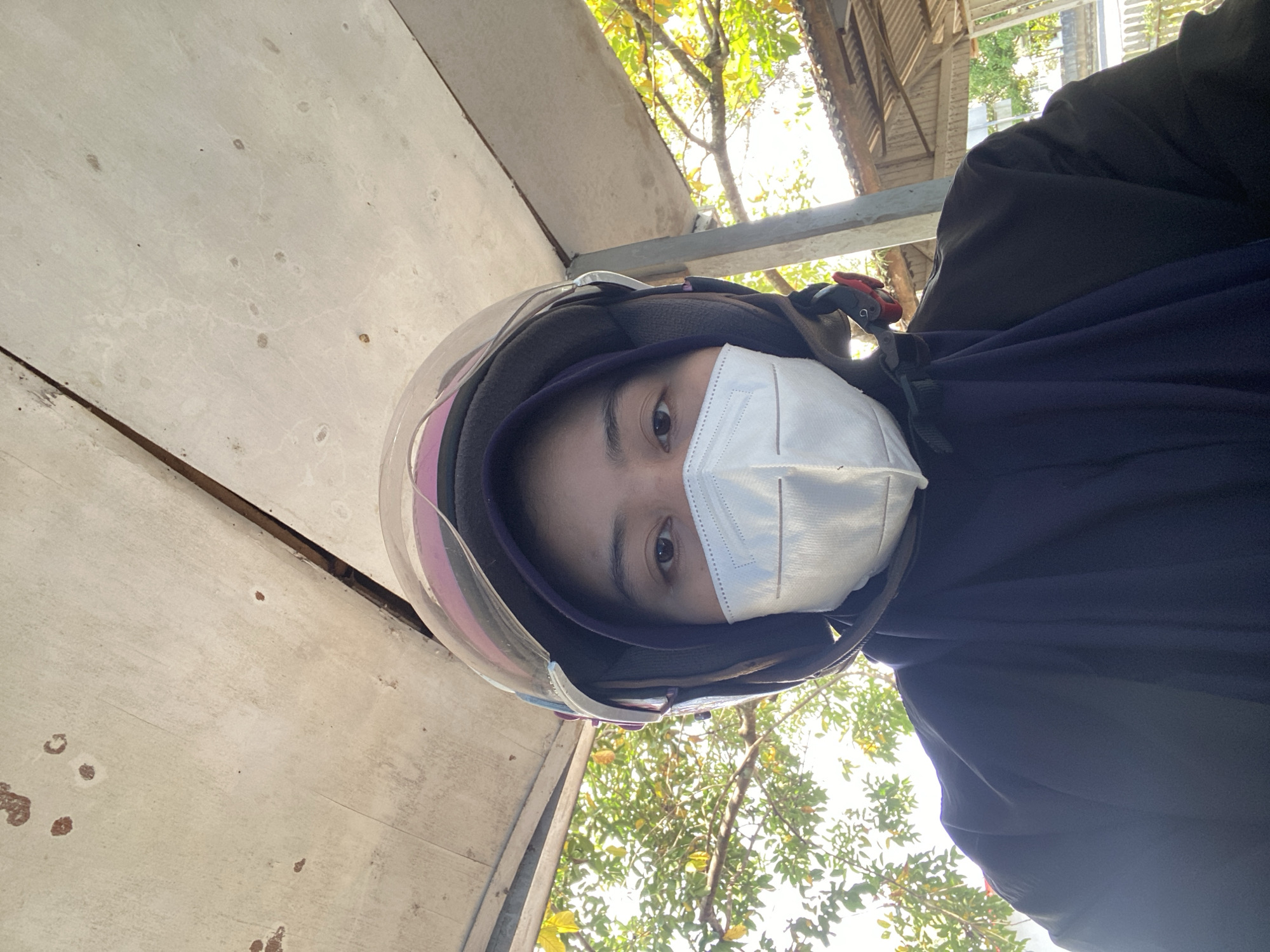A person wearing a dark blue hijab and a white face mask is taking a selfie. They are also wearing a motorcycle helmet with a clear visor. The person is looking directly at the camera. The background shows green trees and foliage, suggesting an outdoor environment. A corrugated metal roof structure is visible in the upper right portion of the image. The overall lighting indicates daytime. The image appears to have been taken from a low angle.