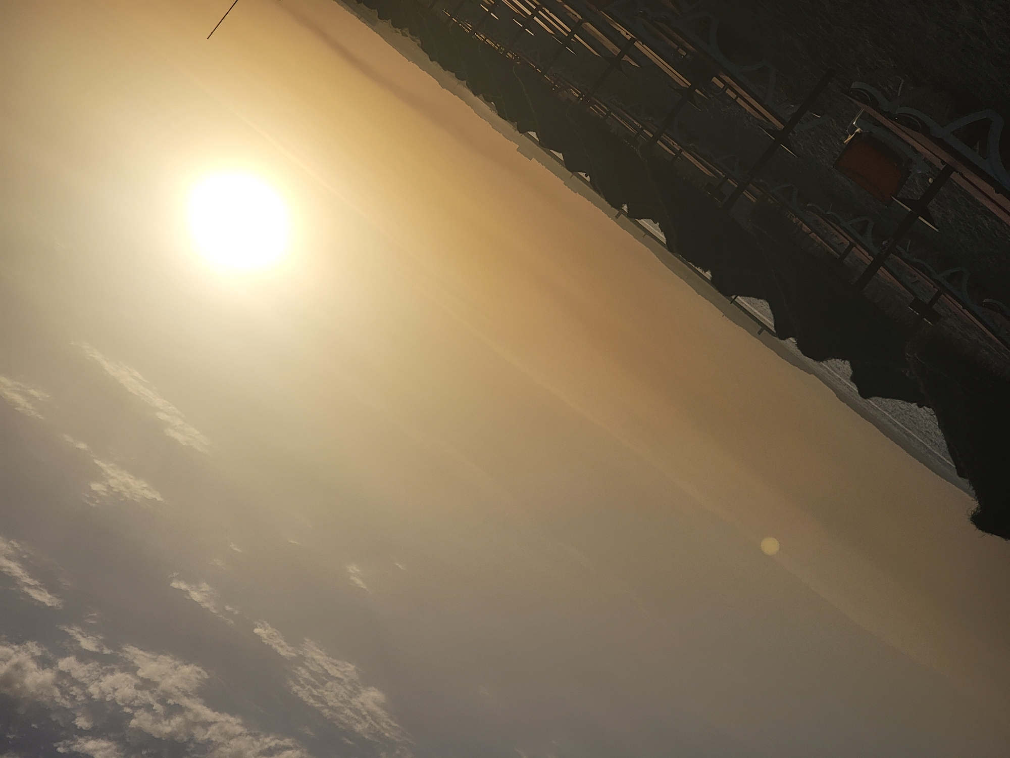 The image depicts a beach scene in Quarteira, Portugal, during what appears to be sunset or sunrise. The sun, a bright, glowing orb, is positioned in the upper right quadrant of the frame, casting a warm, golden light across the sky. Wispy clouds are scattered across the sky. In the foreground, rows of empty sun loungers and umbrellas are visible, suggesting a closed or quiet beach. The dark silhouettes of the loungers and umbrellas contrast with the bright sky. A dark, rocky formation is present in the lower left of the image, possibly a natural feature of the coastline. The scene is captured at an angle, with the horizon line running diagonally from the bottom left to the top right. The overall impression is one of tranquility and the end of a day at the beach.