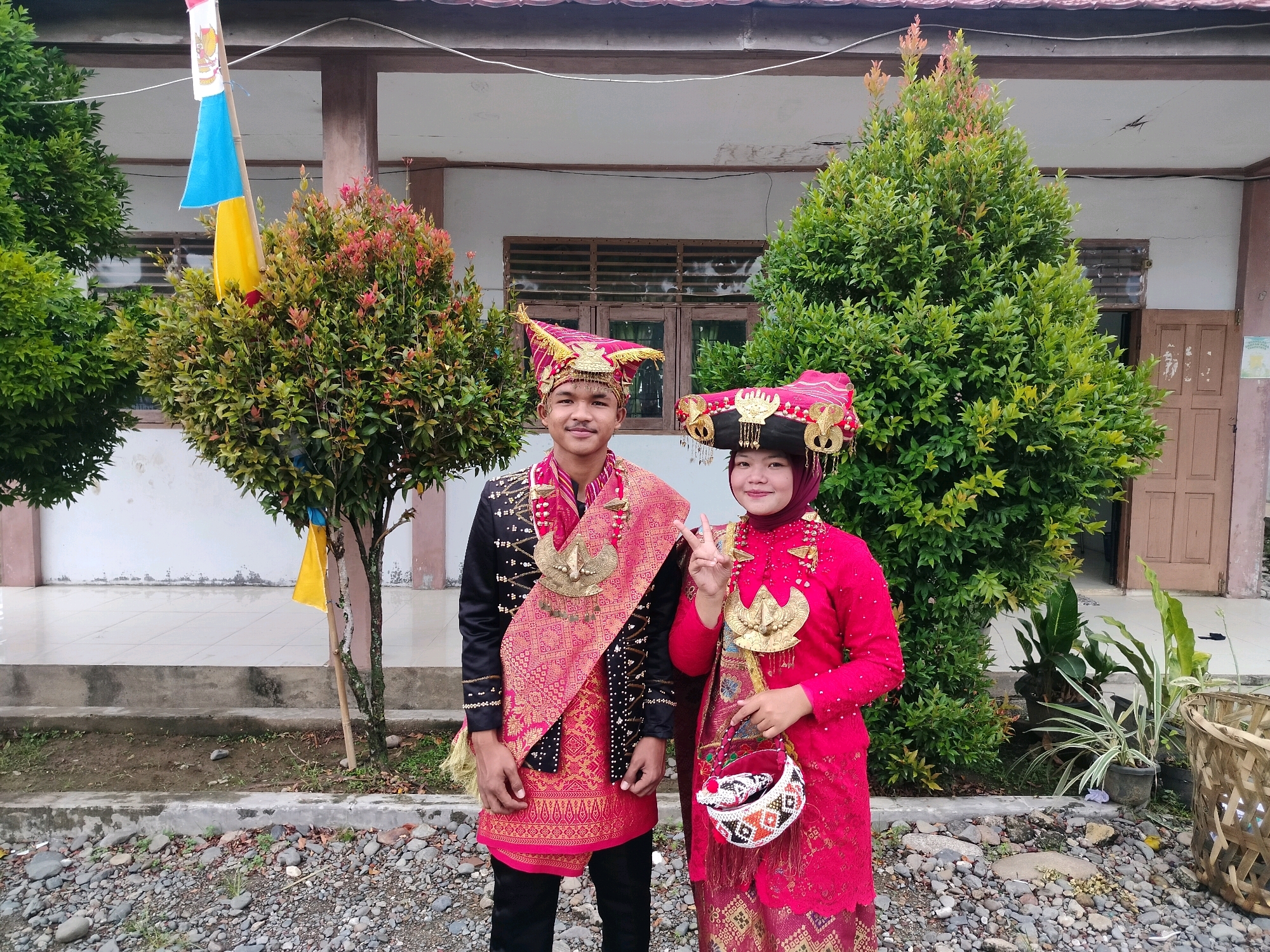 Two individuals, a male and a female, stand side-by-side outdoors, posing for a photograph in front of a building with wooden doors and windows, surrounded by greenery and a gravel-covered ground. The building appears to be a school or community center, given the flags displayed on a pole to the left of the frame, one of which is a red, white, and blue Indonesian flag. The male is dressed in a traditional ceremonial outfit consisting of a black long-sleeved shirt adorned with gold and red patterns, a red sash with intricate gold embroidery, and a matching red headdress. He is also wearing black pants. The female is wearing a vibrant pink traditional outfit, including a long-sleeved top with ornate gold embellishments and matching patterned pants. She has a pink headscarf and a decorative headdress. She is holding a small, woven bag and making a peace sign with her right hand. Both individuals are adorned with large, gold, bird-shaped pendants. The environment suggests a festive occasion, possibly a cultural event or celebration in Lawebekung, Indonesia.