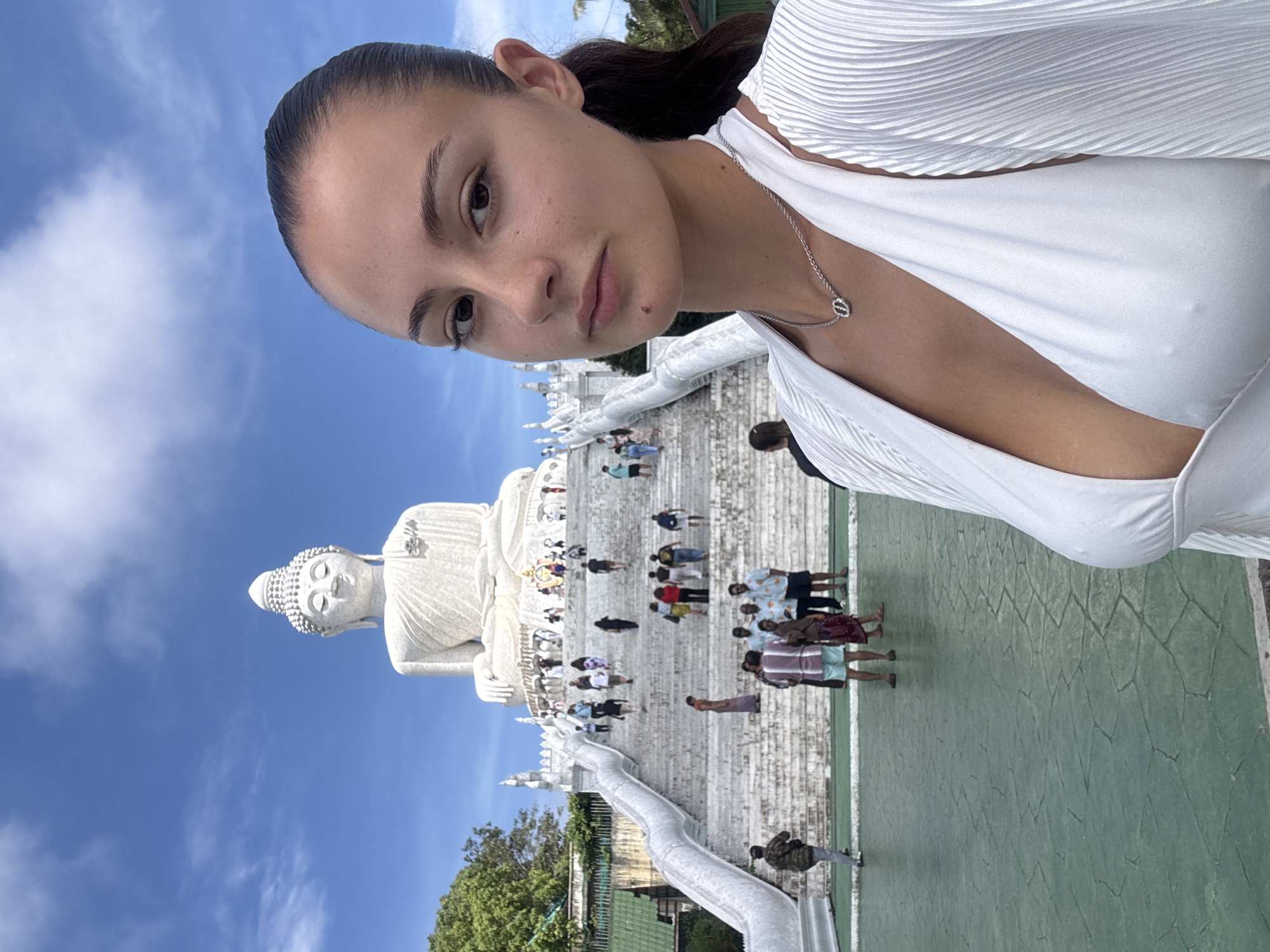 A woman is taking a selfie in front of the Phuket Big Buddha in Karon, Thailand. The statue, made of white marble, is seated atop a hill. Numerous tourists are visible on the stairs leading up to the statue. The woman wears a white top and a silver necklace. The sky is blue with some clouds.