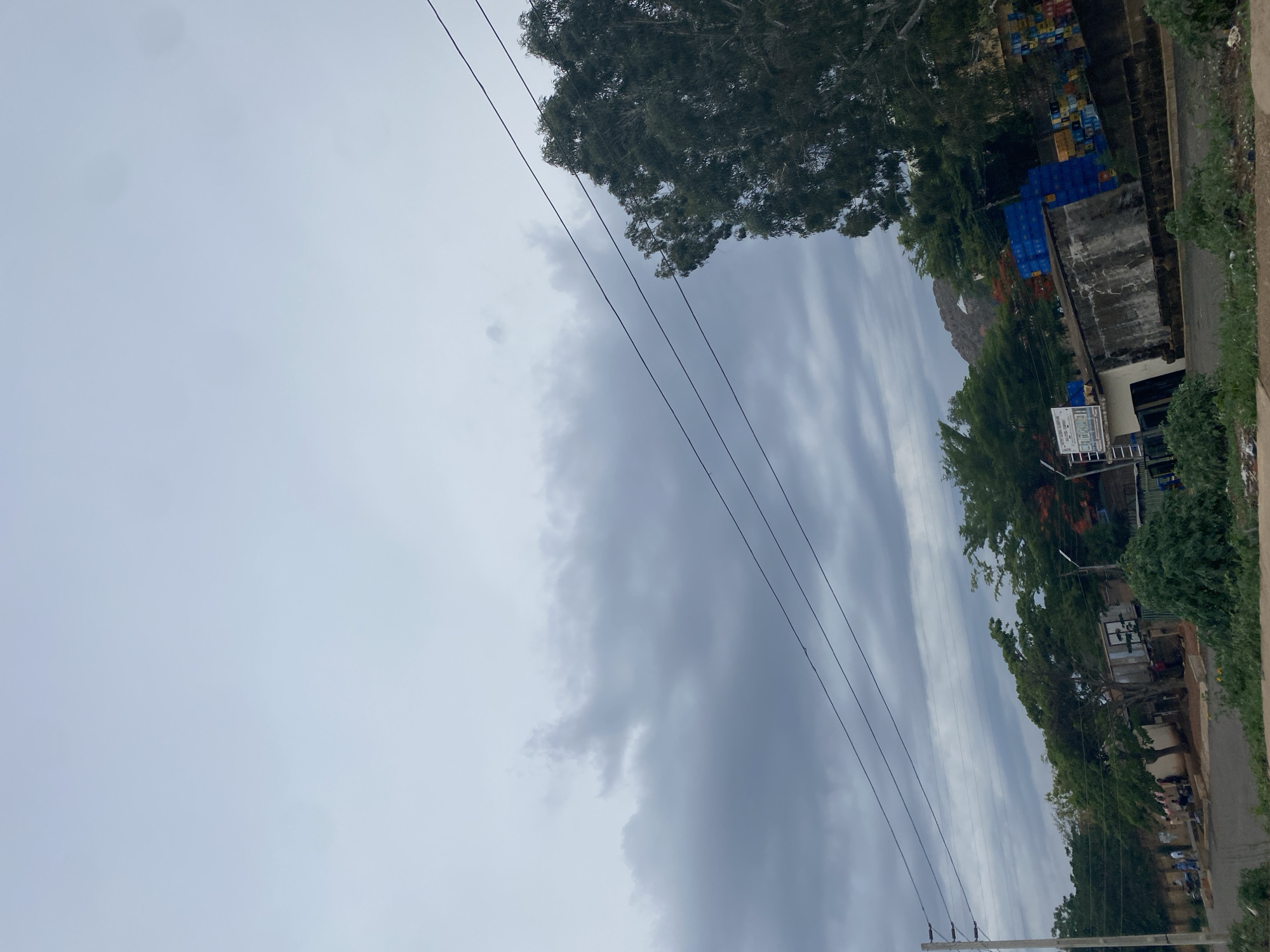 The image shows an overcast day in what appears to be Gura Top, Nigeria. Overhead power lines run diagonally across the frame. To the right, a road with some vegetation alongside it is visible. Adjacent to the road is a building with blue crates stacked behind it. Trees are interspersed along the roadside and behind the building. The sky is predominantly gray with cloud formations. There is no visible human activity.