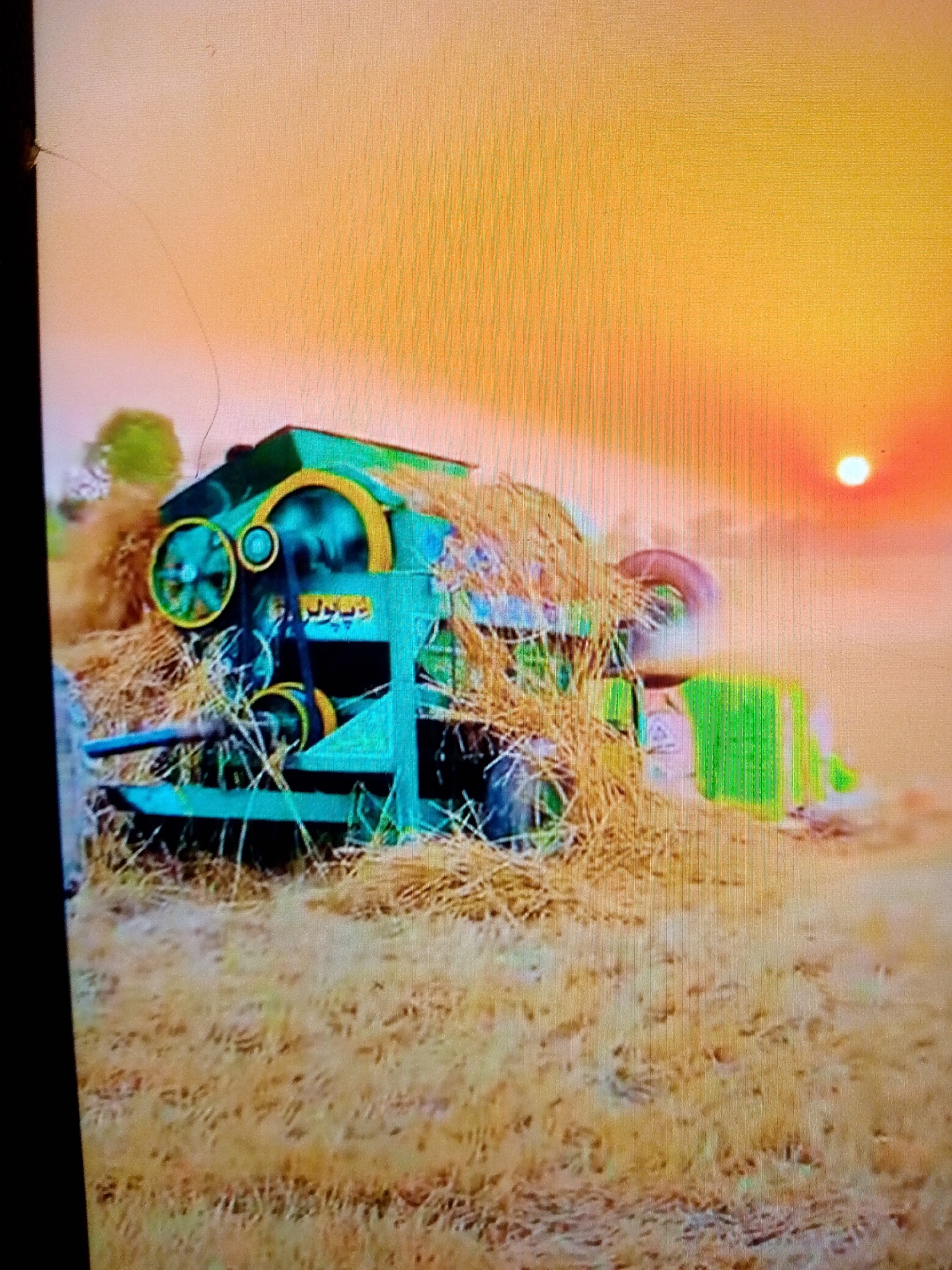 The image depicts an agricultural scene featuring a green and yellow thresher machine actively processing harvested crops in an open field. Piles of dry straw and chaff surround and partially cover the machine, indicating its operation in separating grain. The thresher has prominent yellow-painted wheels, belts, and an arched structure. On its side, yellow text is visible, which appears to be "بیوپاری" (Biopari) in Urdu/Punjabi script.
The setting is an agricultural field with light brown, dry crop residue on the ground. The time of day is late afternoon or early evening, characterized by a vivid orange and yellow sky that dominates the upper right portion of the frame. A bright, spherical sun is clearly visible as a glowing white disc against the warm sky, suggesting sunset. The weather appears clear and calm, with soft, warm light illuminating the scene. No people are visible. The location context for this rural activity is Karamsar, Pakistan. The image displays some digital artifacts, likely from being a photograph of a screen.