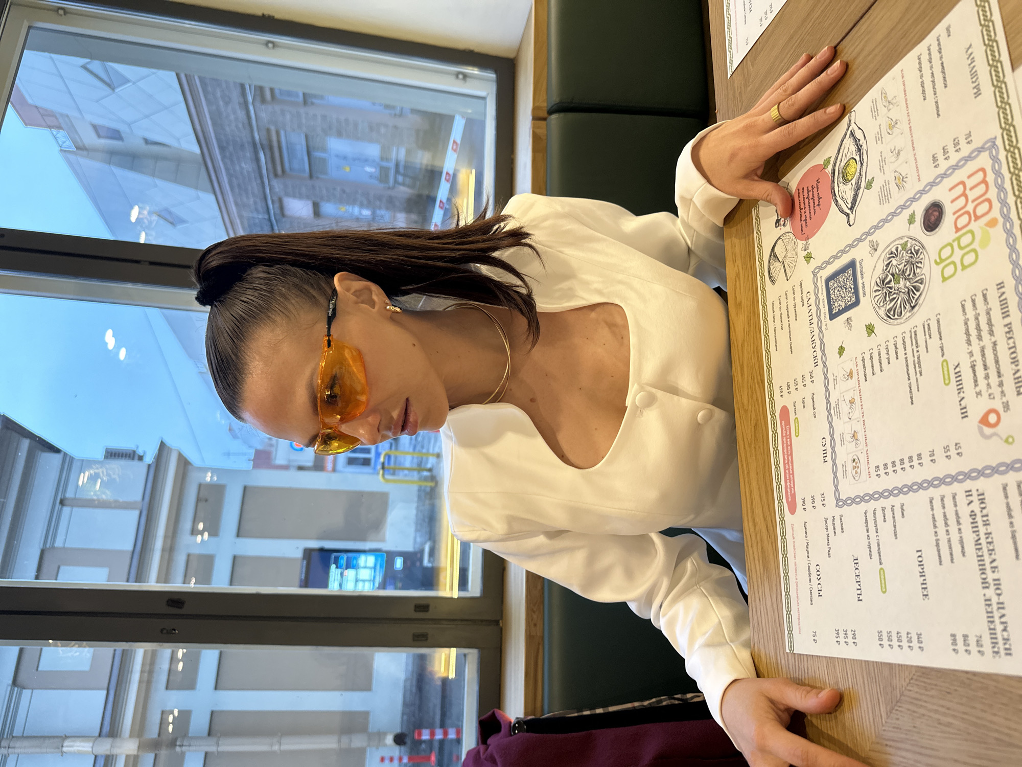 A medium shot, oriented vertically, shows a woman sitting at a table in what appears to be a restaurant. The restaurant's menu is visible on the table. The woman wears orange sunglasses and a white top. Outside the window, buildings are visible, indicating a setting in Saint Petersburg, Russia. The menu, printed in Russian, lists various dishes, including khachapuri, kebabs, and salads. The restaurant promotes the Saint Petersburg locations, on Nevsky avenue, Moscow avenue, and Yefimova street.